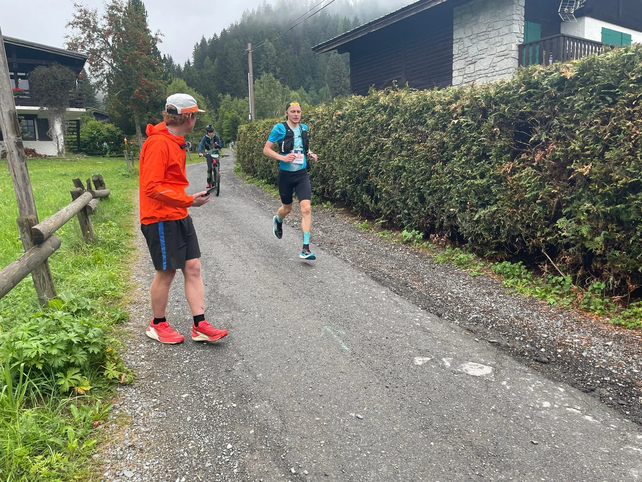 A runner going through a mountain village, followed by a mountain biker, while the coach is looking and giving advice