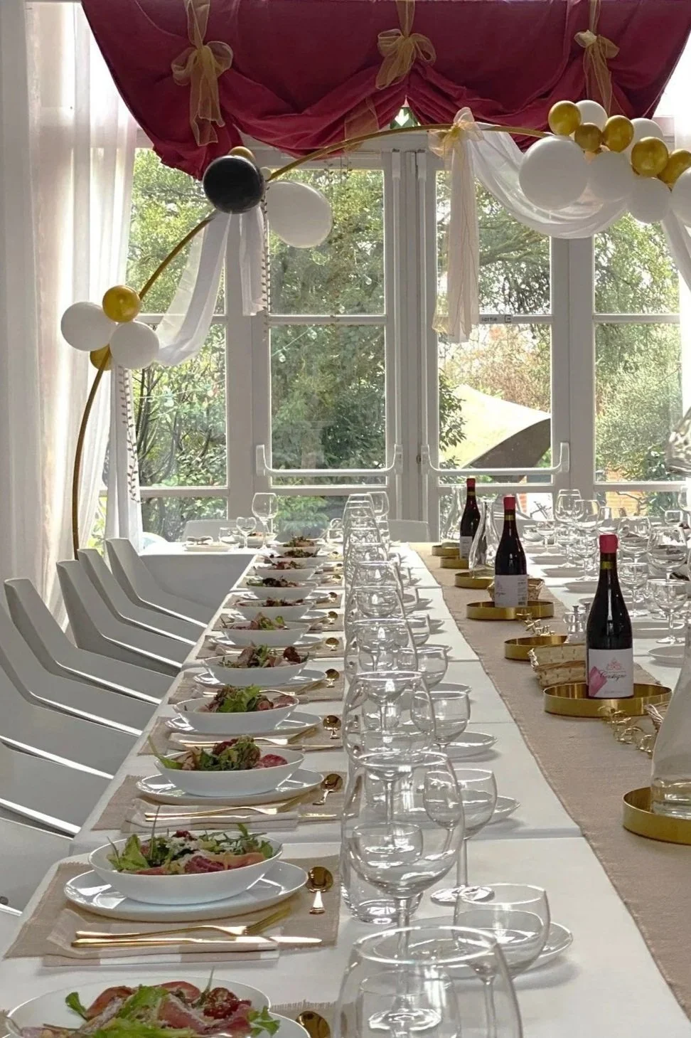 Table dressée pour un repas de fête avec assiettes, verres, bouteilles de vin, et arche de ballons et rubans dans une salle lumineuse avec fenêtres donnant sur des arbres.