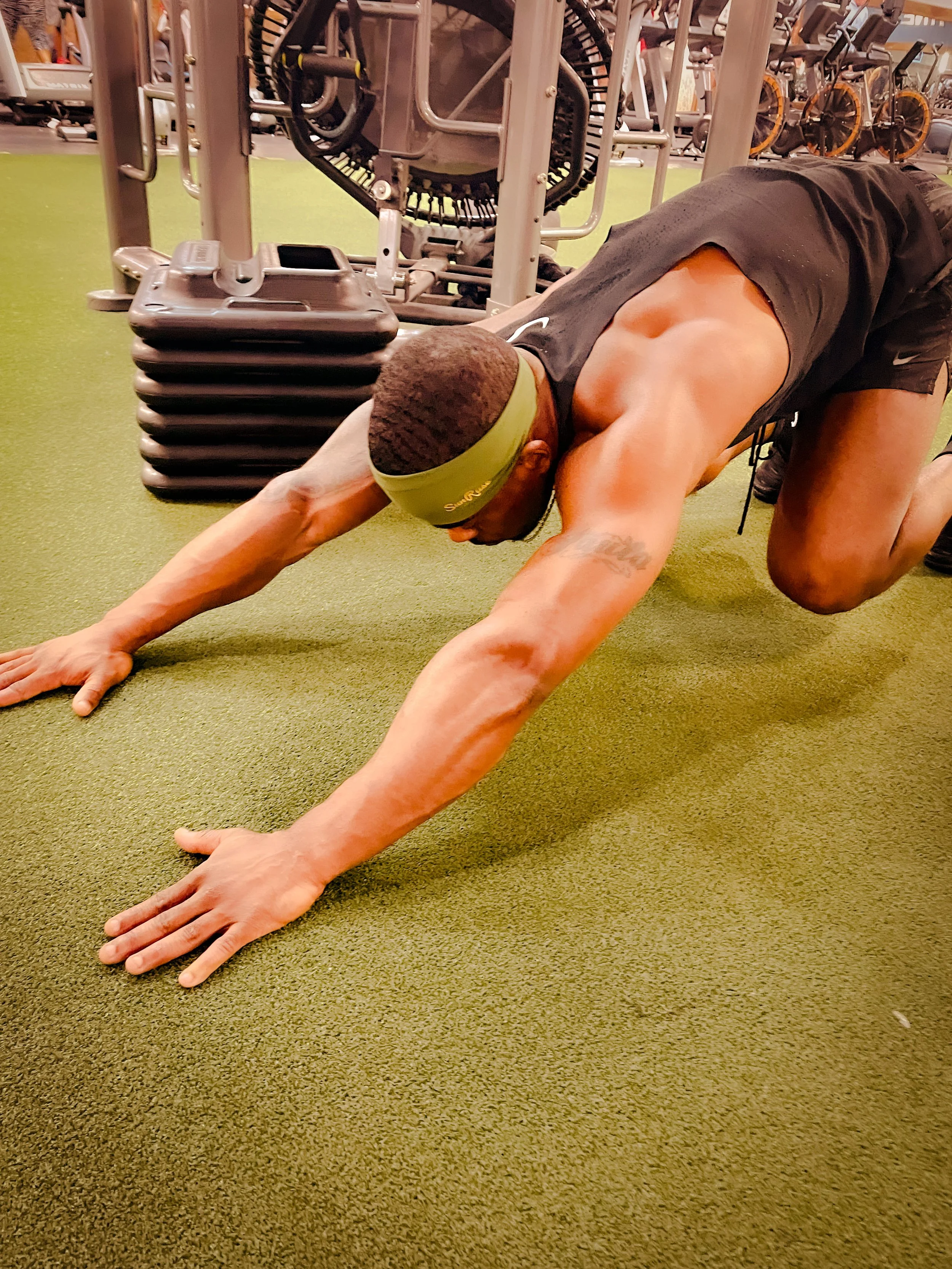 A man doing a plank exercise on a green gym floor, wearing a green headband, black tank top, and black shorts, in a gym with various workout equipment in the background.