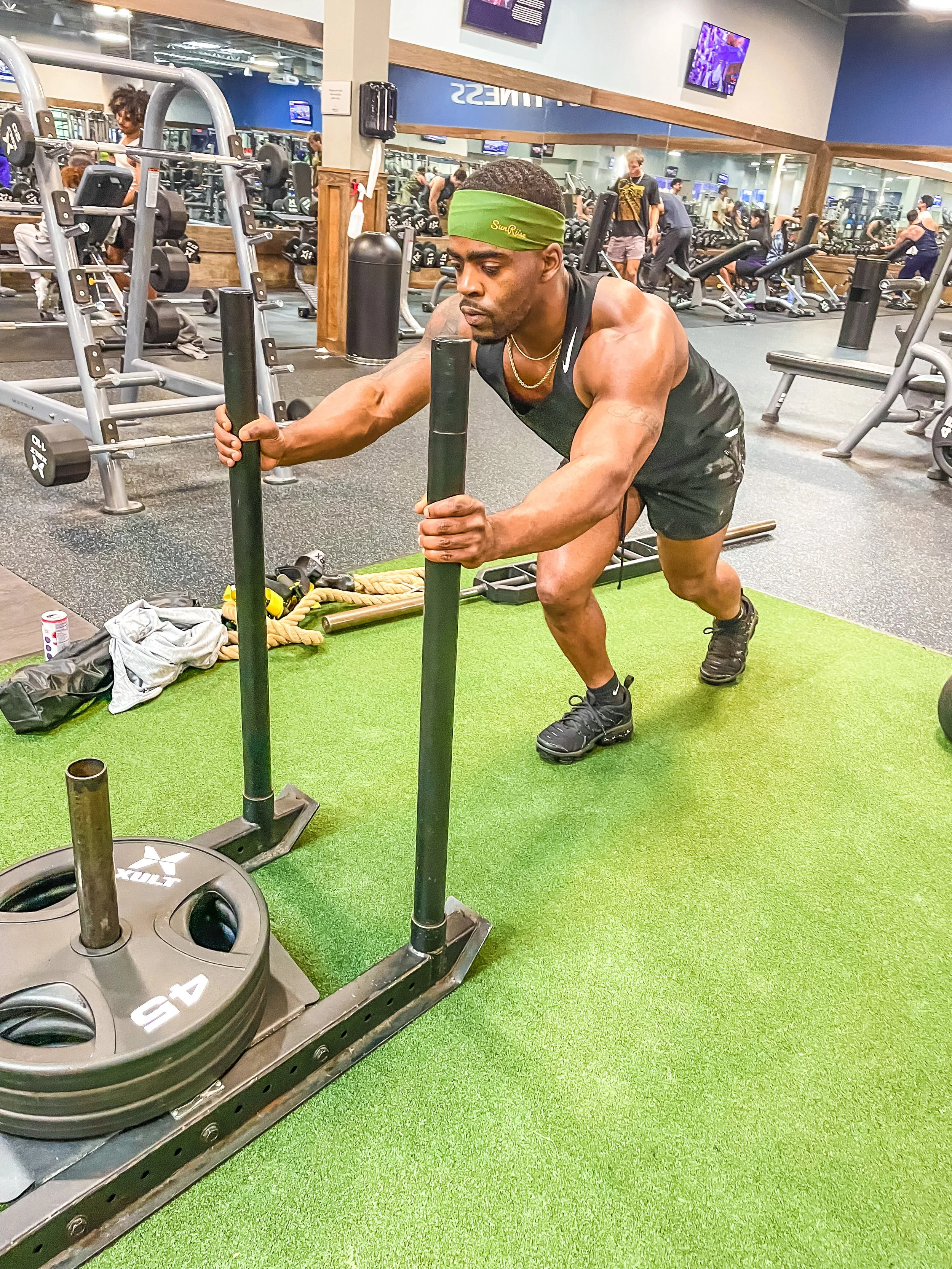 A man in a gym pushing a weighted sled on a green turf floor, surrounded by gym equipment and other people working out in the background.