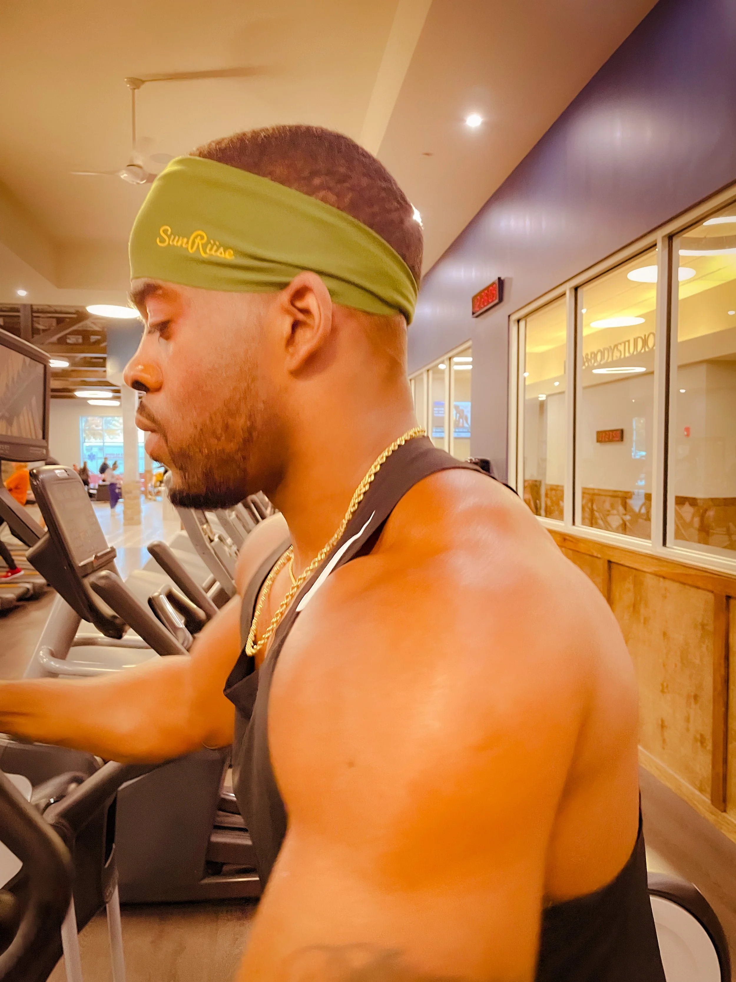 A young man with a green headband, gold chain, wearing a black tank top, working out on gym equipment in a fitness center.