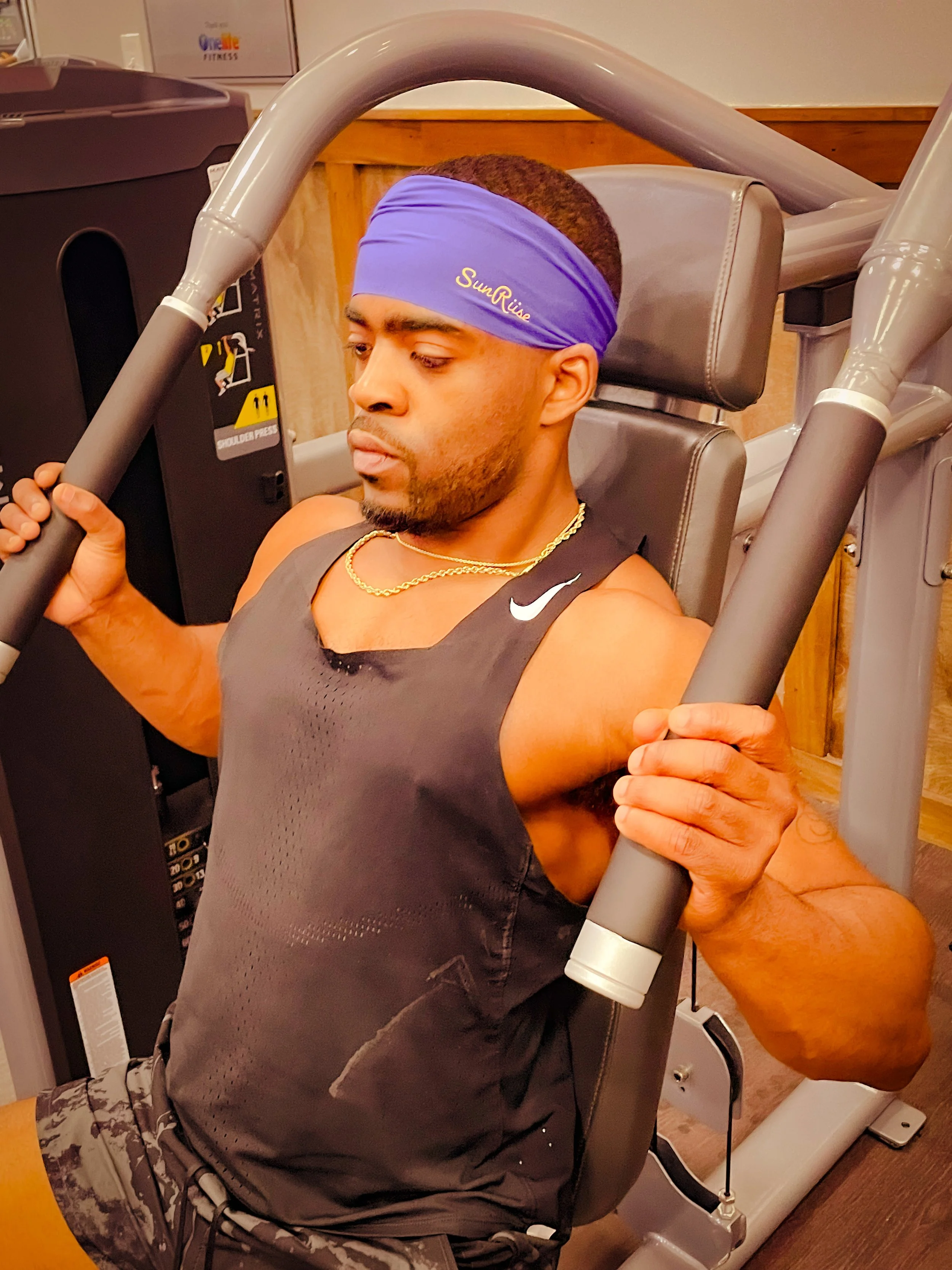 Man working out on a shoulder press machine in a gym, wearing a purple headband, black athletic tank top, camouflage shorts, and gold chains.