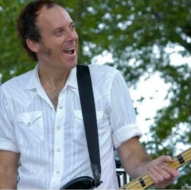 A man playing an electric guitar outdoors, wearing a white short-sleeved shirt and smiling.