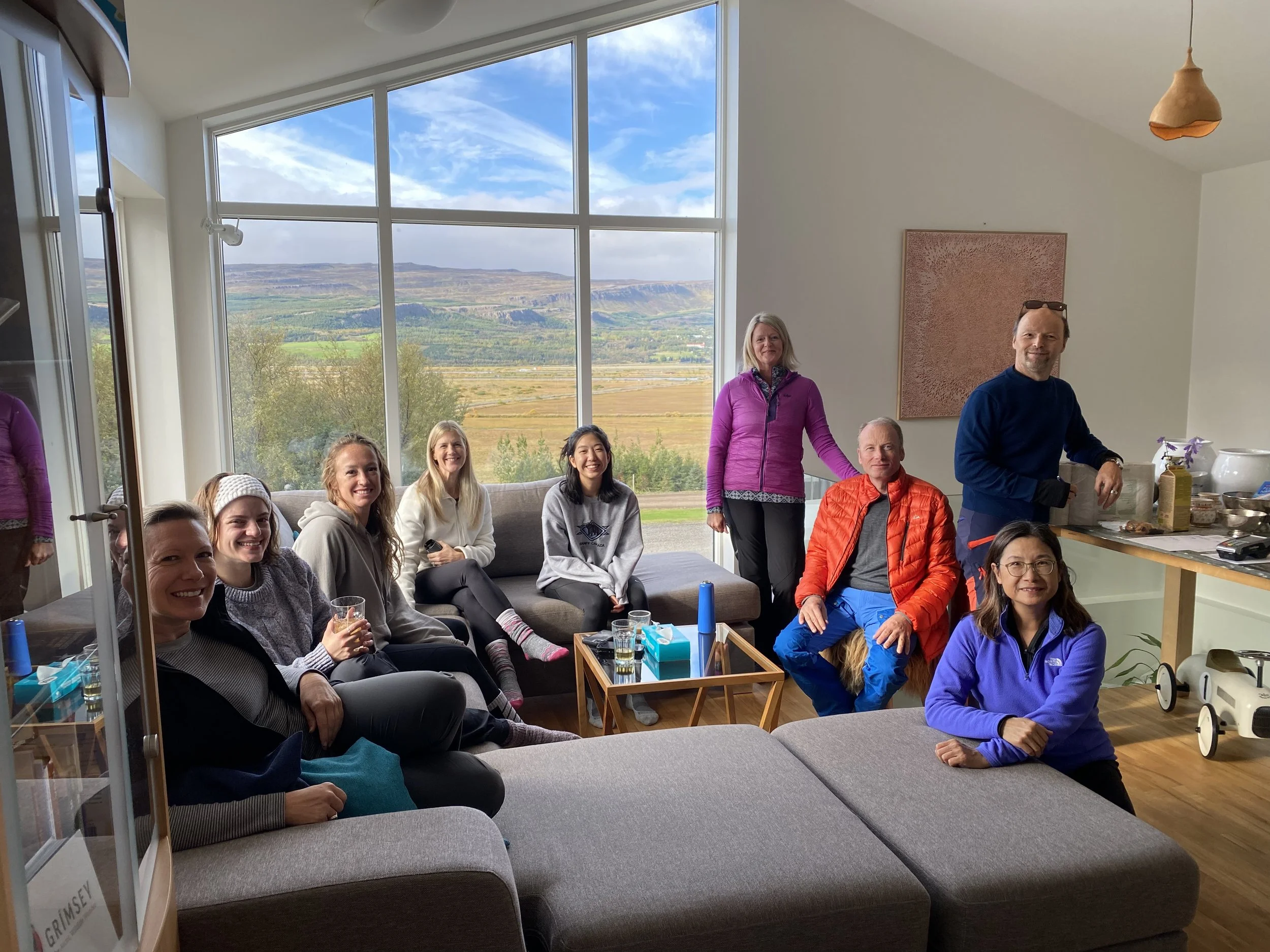 Group of people sitting in a living room with large windows showing a scenic outdoor view.