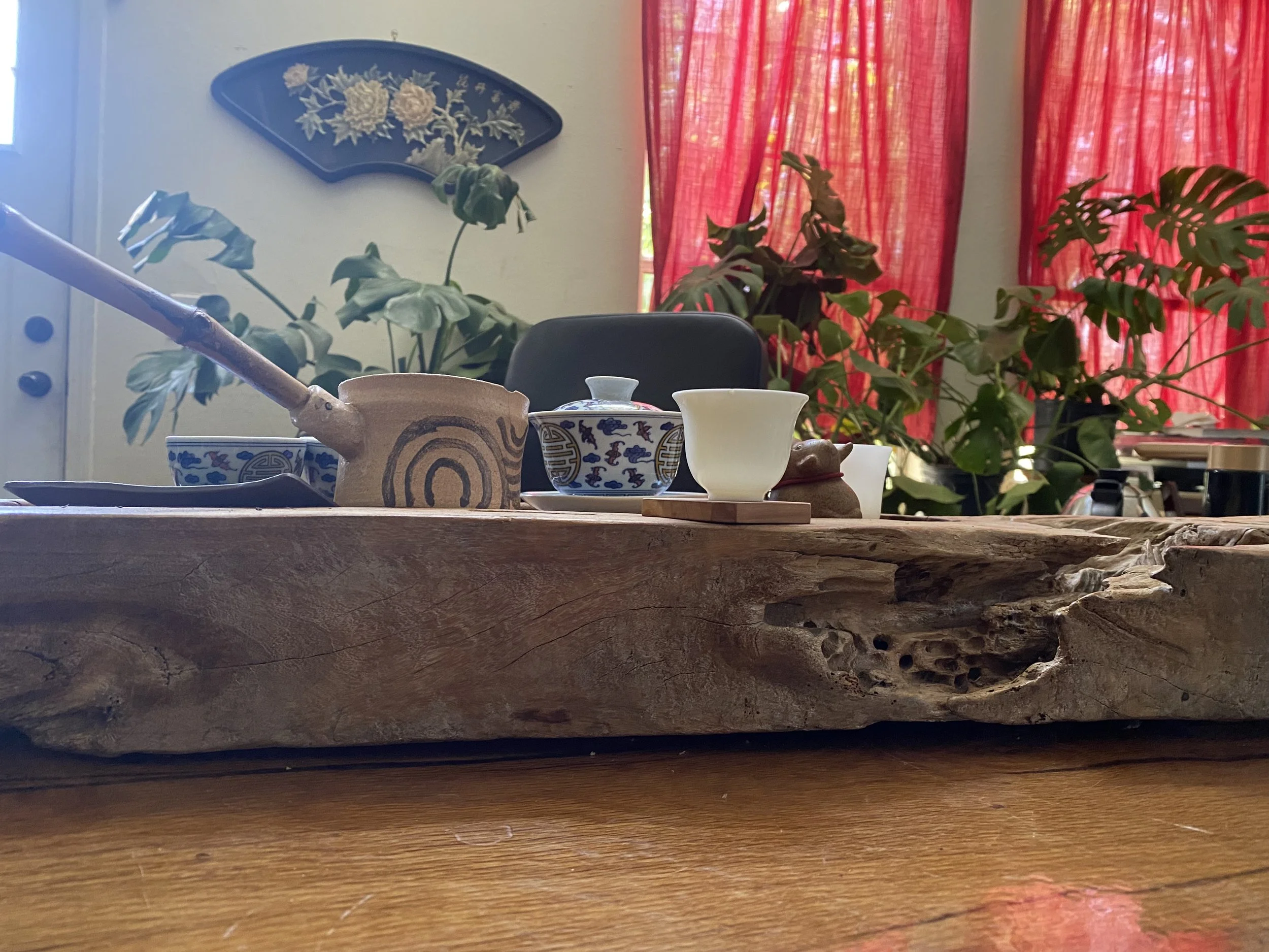 Japanese tea set with teacups on a wooden stand, surrounded by decorative plants, red curtains, and a wall fan decoration in a room.