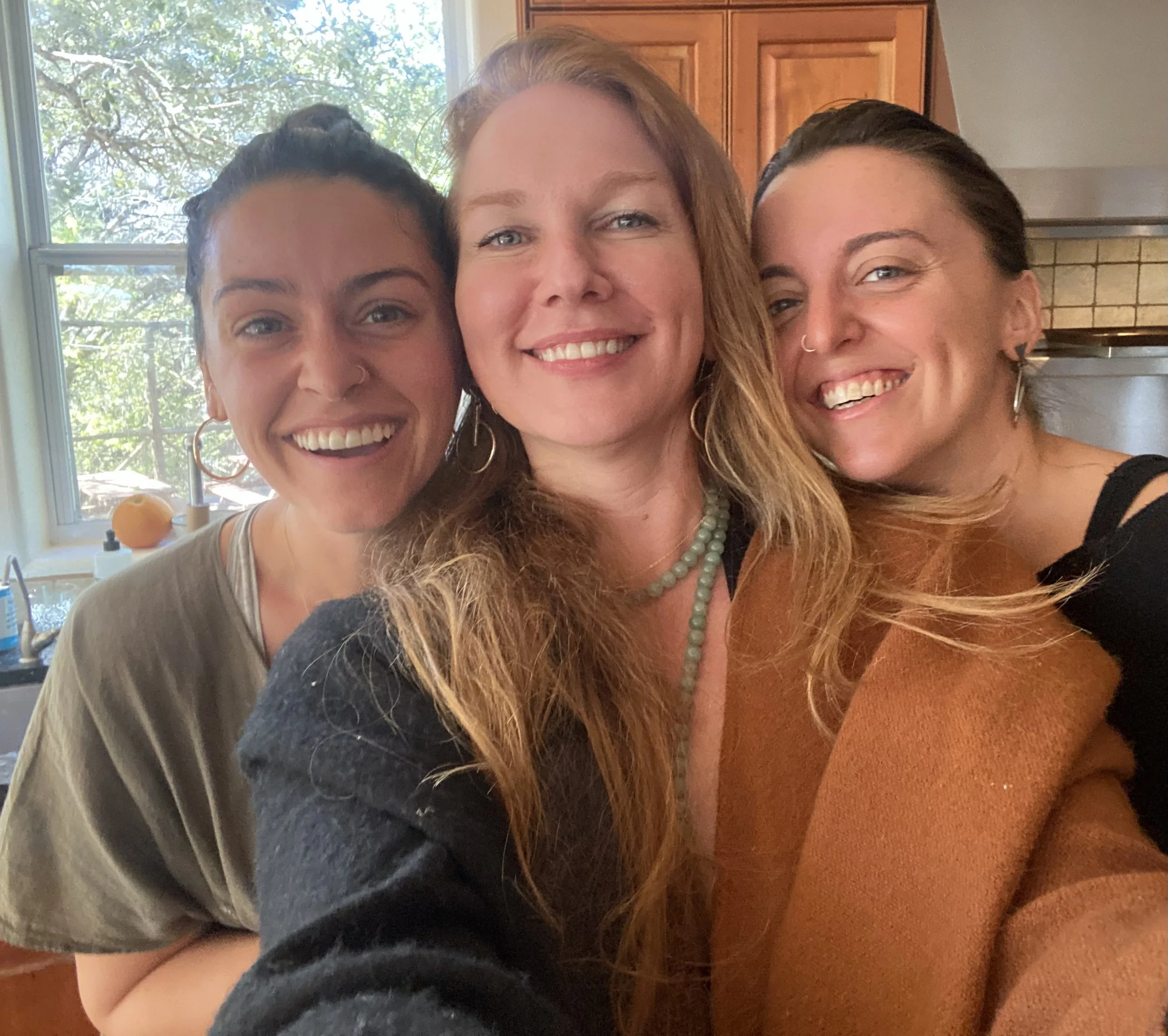 Three people smiling indoors in front of a window and kitchen cabinets.