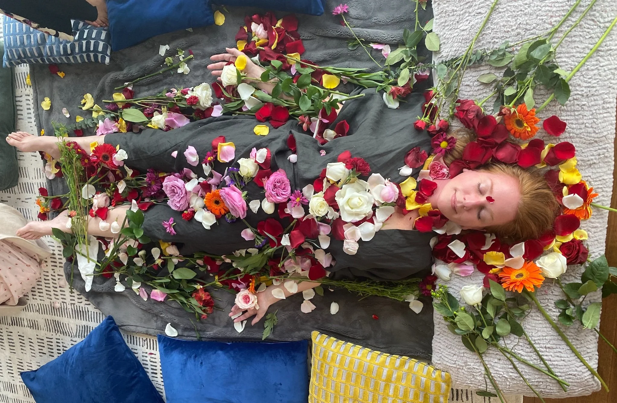 Person lying on a bed of colorful flower petals, surrounded by pillows, appearing serene.