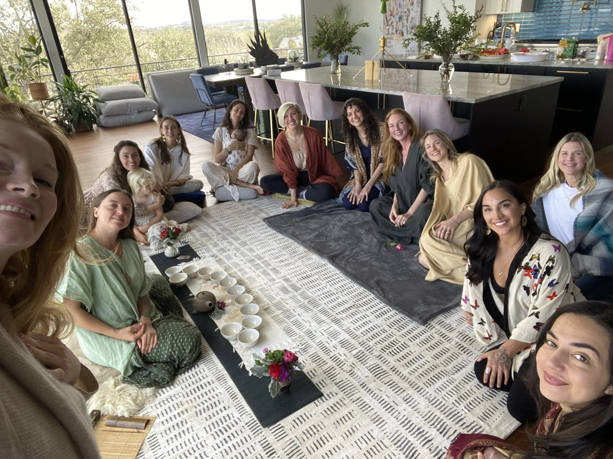 Group of women sitting in a circle on the floor in a modern living room, engaged in a tea ceremony with cups and teapot in the center. The room features large windows and plants.