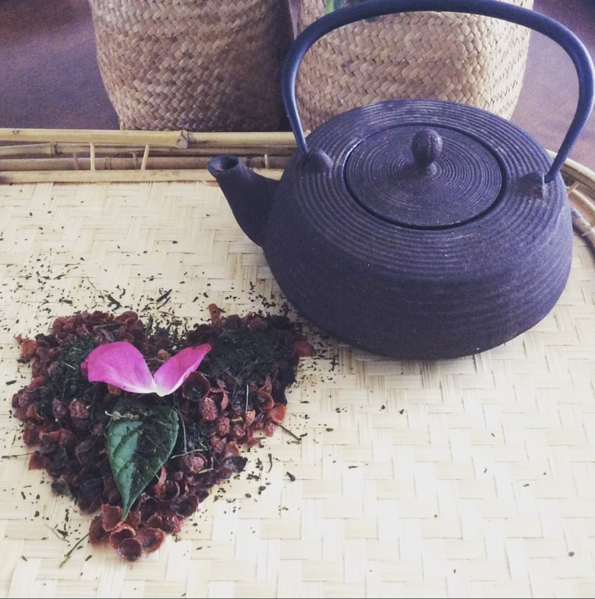 A cast iron teapot on a woven mat beside a heart-shaped arrangement of dried tea leaves and flower petals.