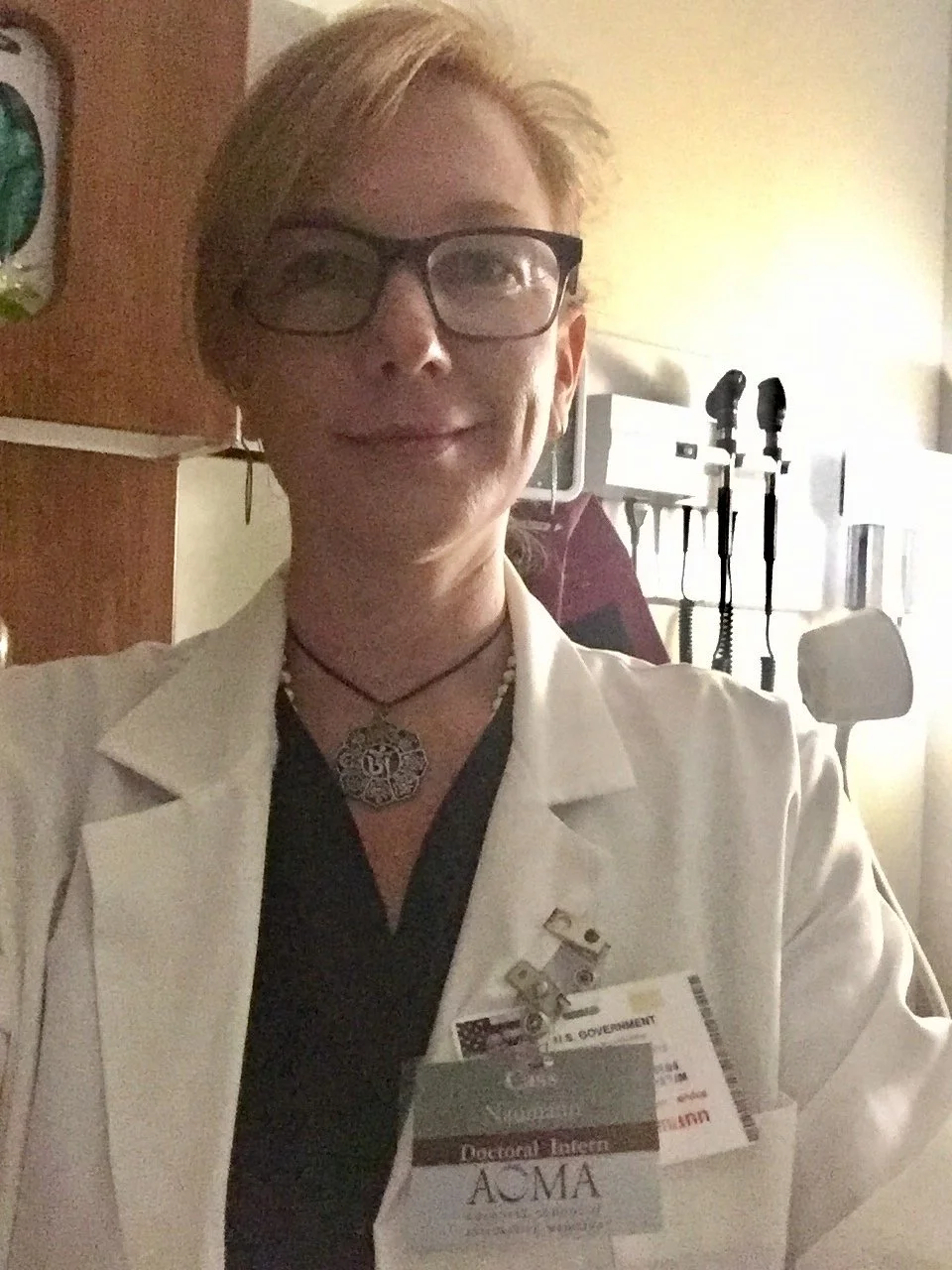 A woman wearing glasses and a white lab coat, with a name badge and decorative necklace, taking a selfie in a medical or hospital setting.