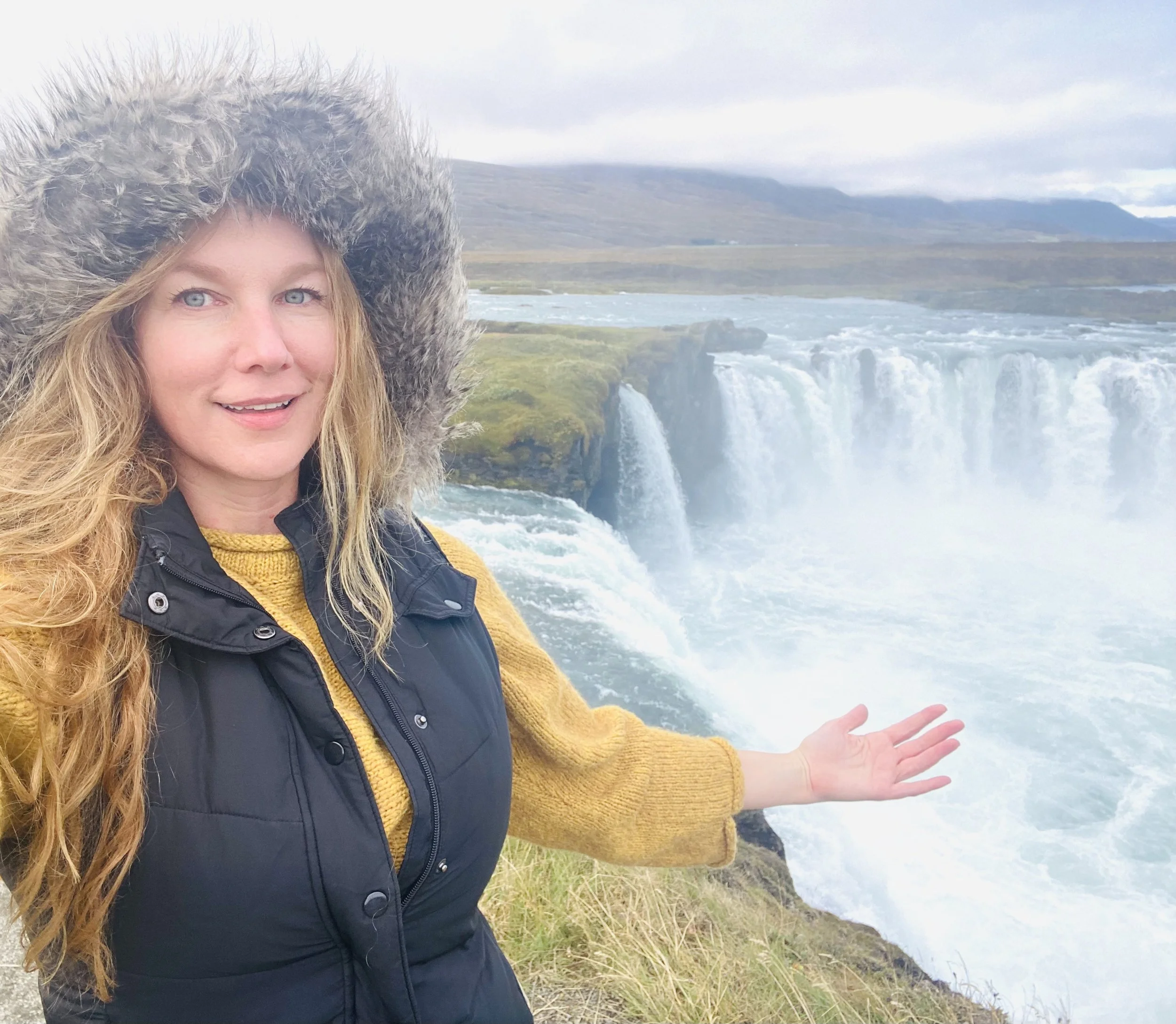 Person in a fur-lined hooded jacket posing near a large waterfall, possibly Godafoss, in a natural landscape with cloudy skies.