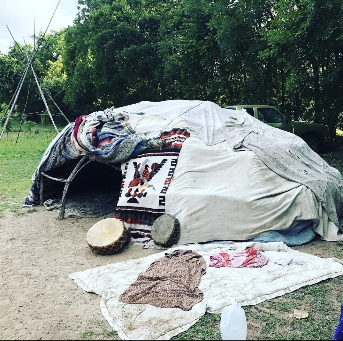 Sweat lodge covered with blankets, drums, and a traditional mat outside; trees in background.
