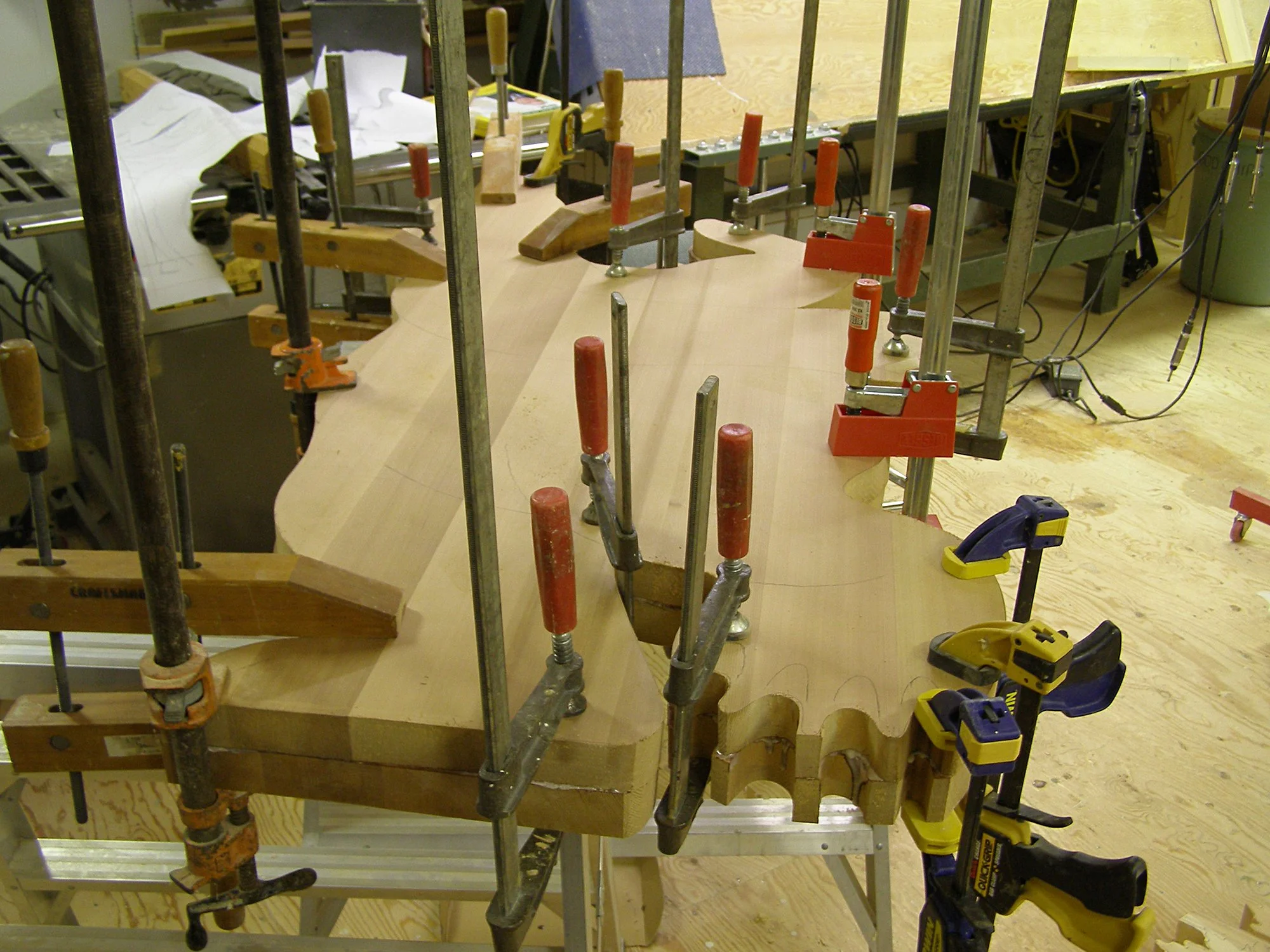 Woodwork project in progress on a workbench, with clamps holding a piece of wood, various tools, and blueprints. A chop saw is visible nearby, and the workspace has a wooden floor and plywood surfaces.