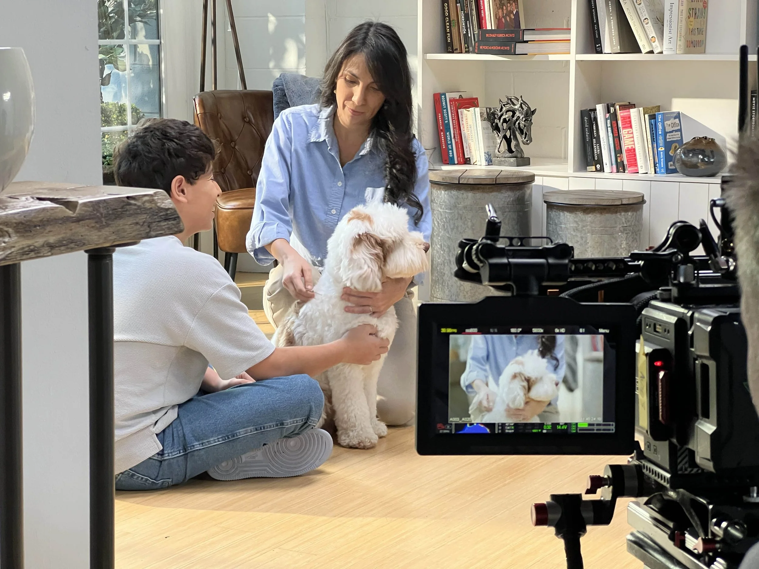 A woman and a boy filming a dog in a living room. The woman holds the dog while the boy touches its paw. A camera records the scene, with a bookshelf and decor in the background.