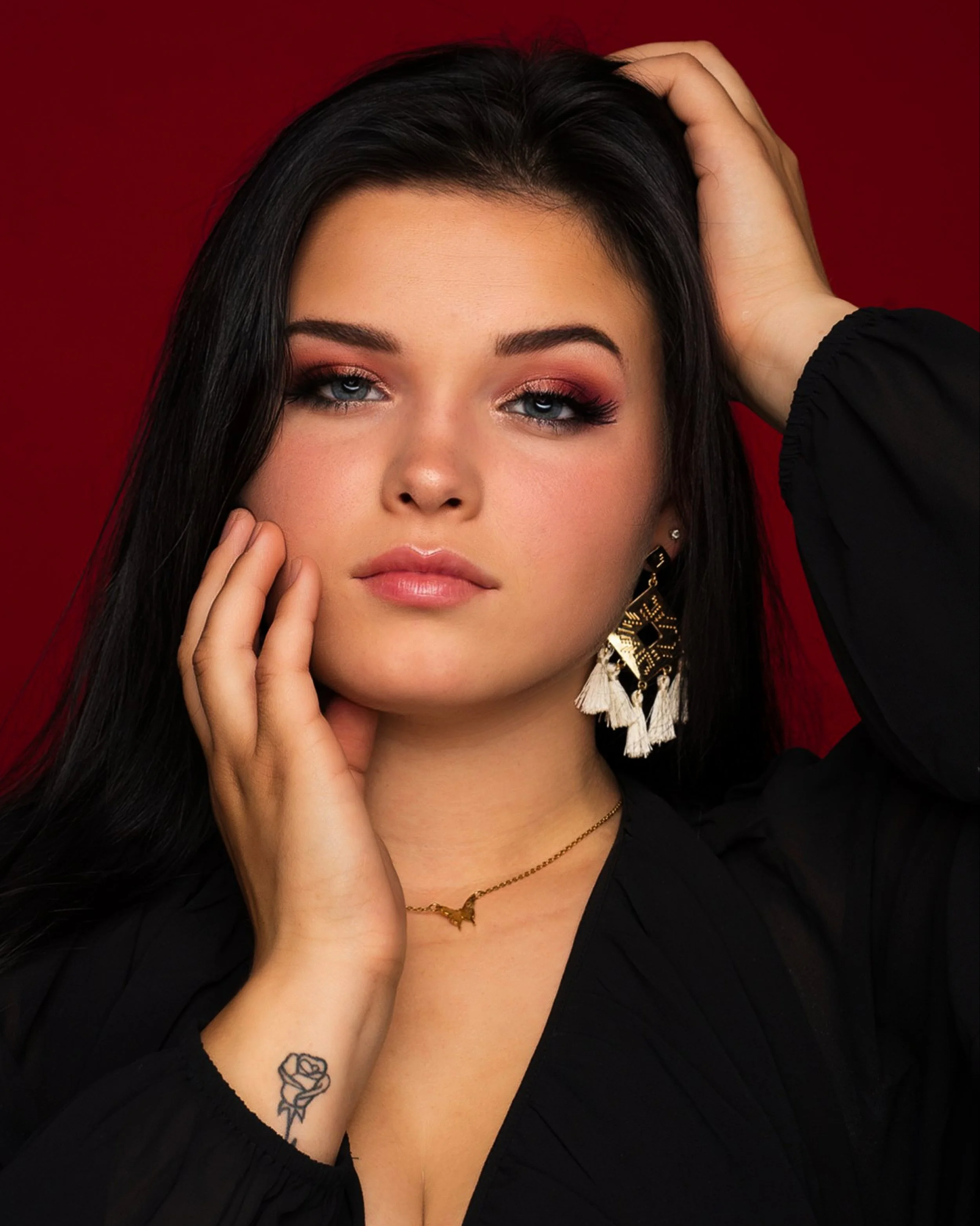 A woman with long dark hair and makeup, wearing earrings, a necklace, and a tattoo of a rose on her wrist, touching her face and head, with a red background.