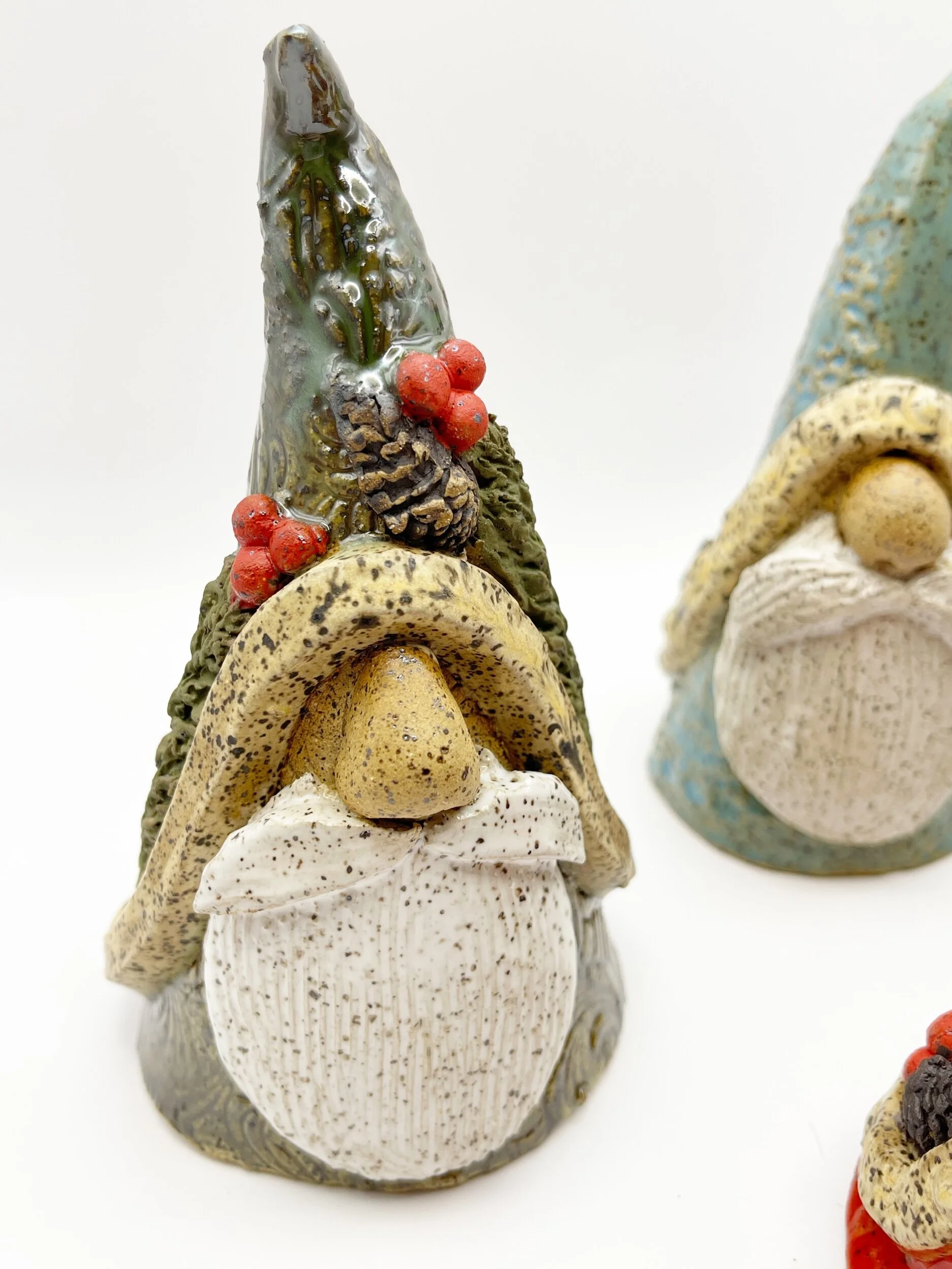 Decorative gnome sculpture with a tall green hat, red berries, pine cone, beige mushroom, miniature gnome face, and speckled white beard, on a white background.