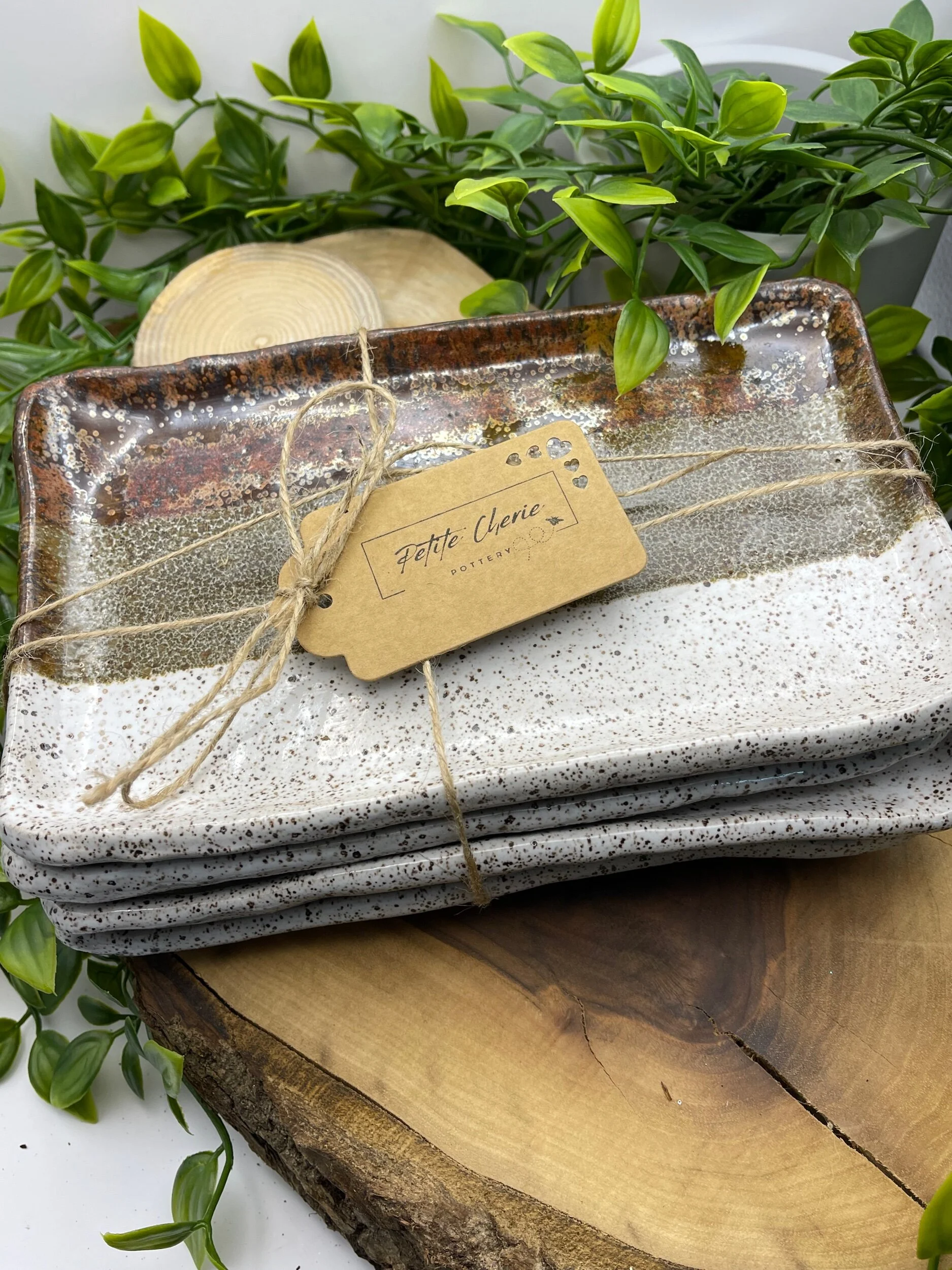 Stack of speckled ceramic trays on a wooden slab, tied with twine, with a brown paper tag labeled 'Feltte Cherie Pottery', surrounded by green plants.