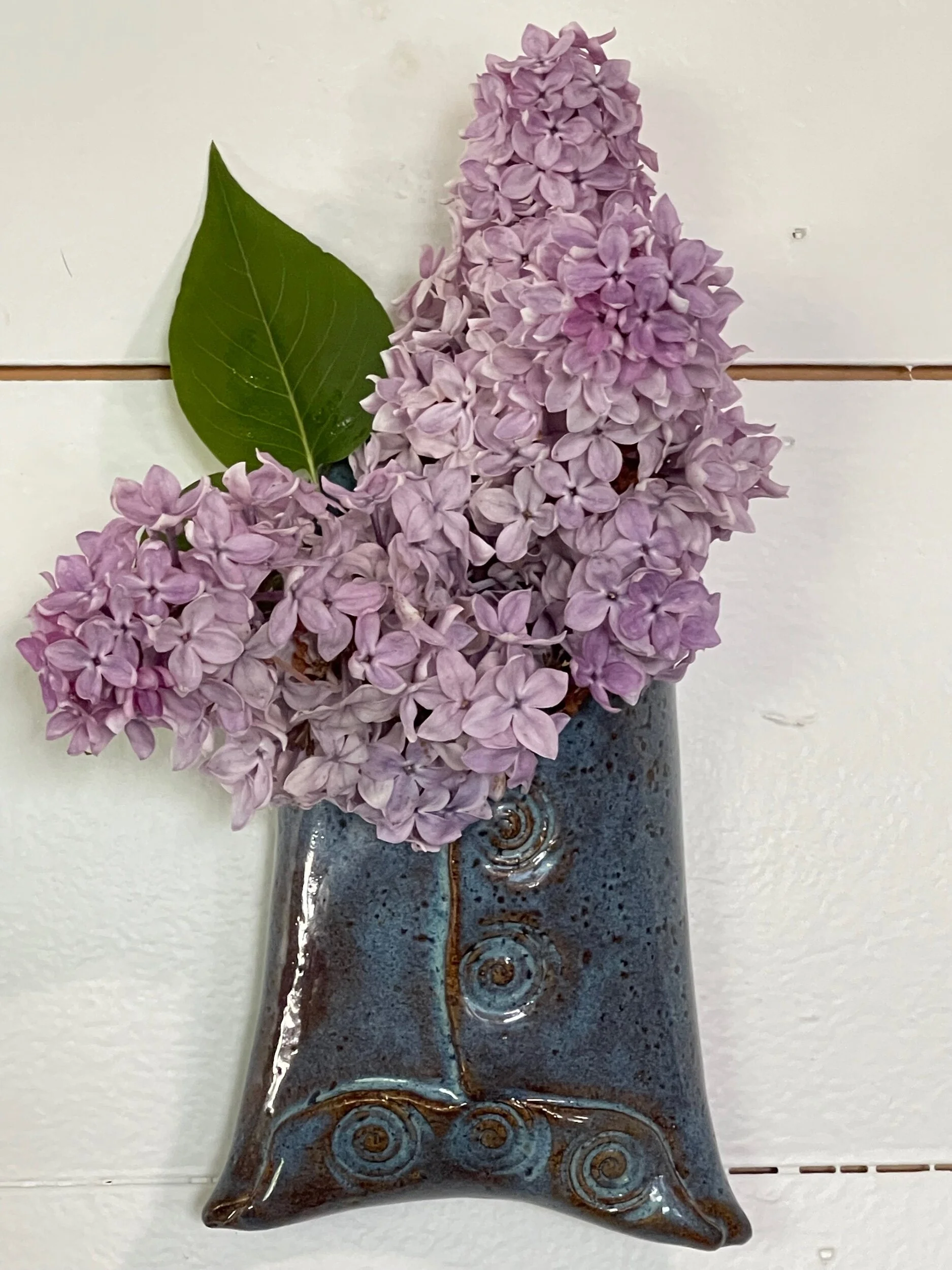 Pink hydrangea flowers in a decorative ceramic vase with a dark glaze, placed on a white tiled surface.
