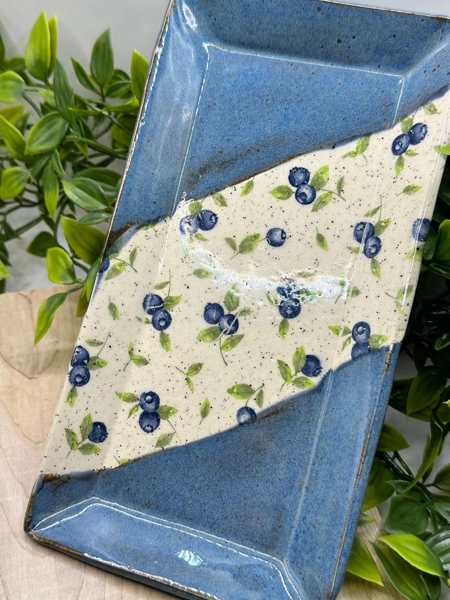 A ceramic rectangular tray with a blue glaze and a white center decorated with small, dark blue blueberries and green leaves. The tray is placed on a wooden surface with some green foliage in the background.