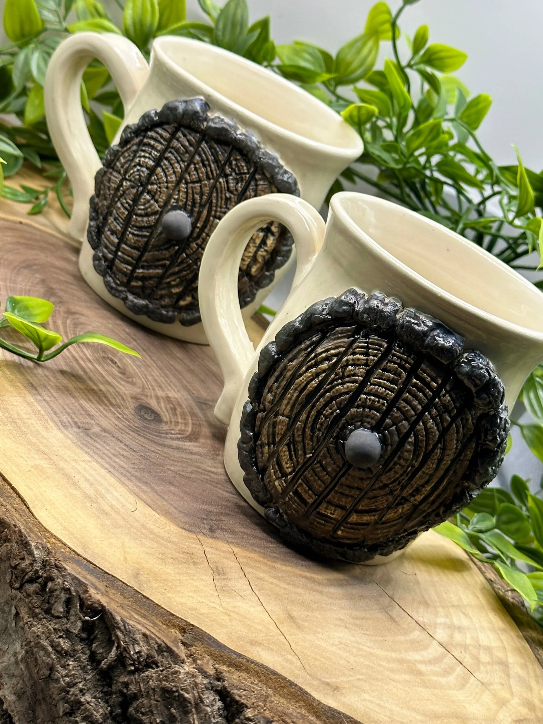 Two beige ceramic mugs with large decorative black and brown circular appliqués on the front, placed on a wooden surface with green leafy plants in the background.