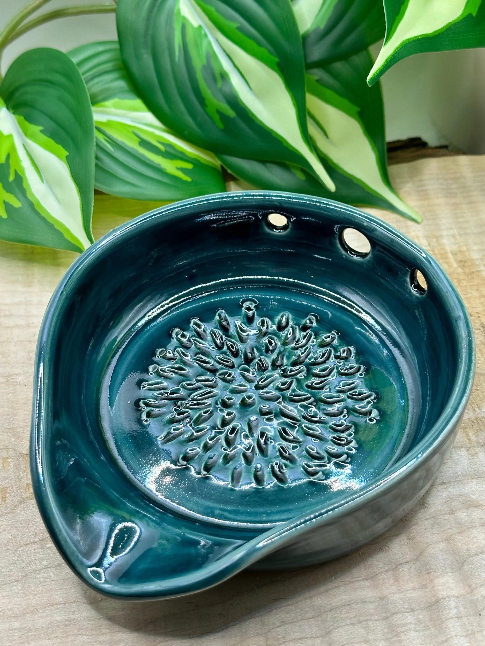 A ceramic soap dish with a glossy teal finish and raised floral design in the center, placed on a wooden surface with green and white variegated leaves in the background.