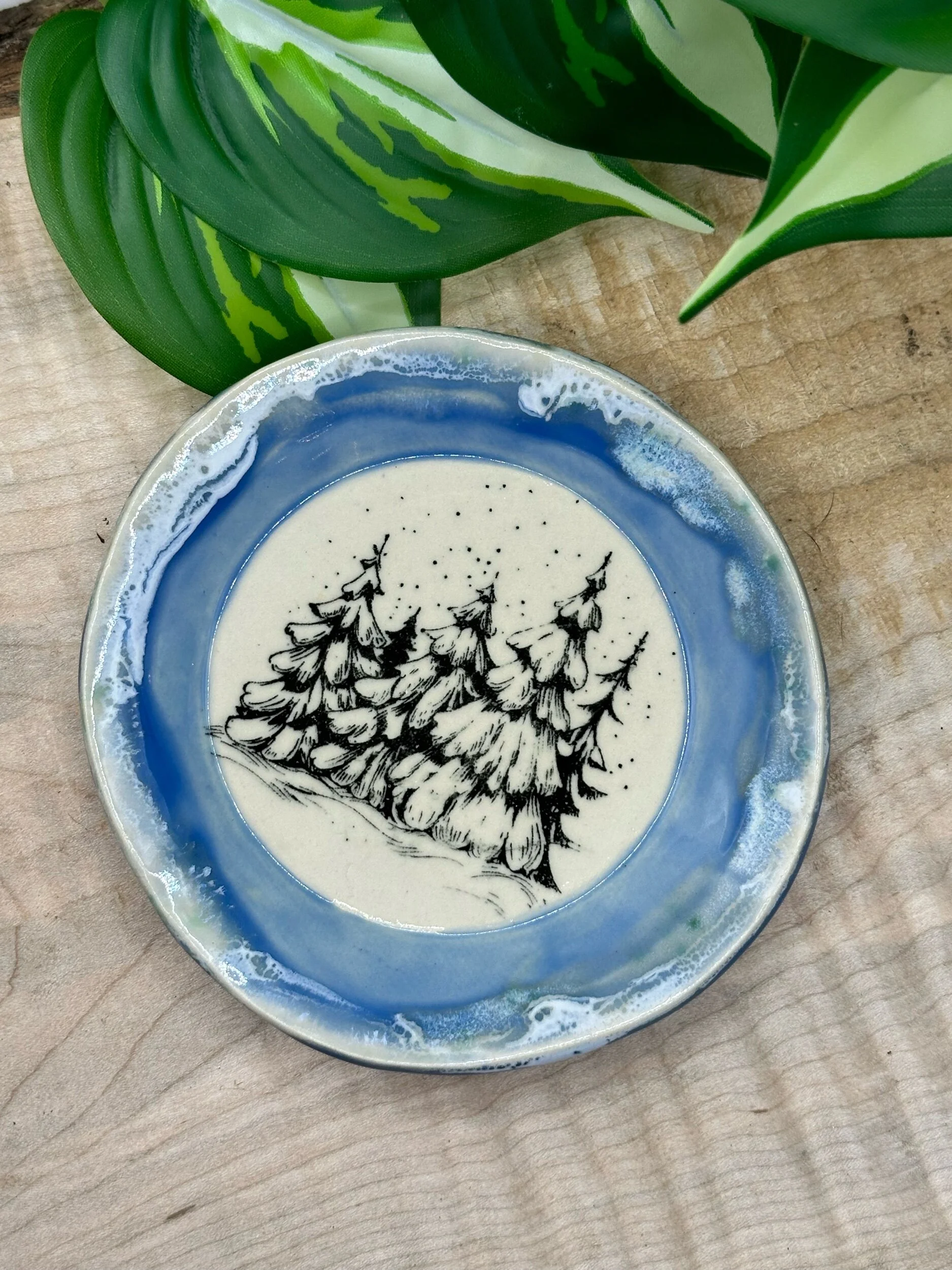 Decorative ceramic dish with a black ink drawing of pine trees on a snowy landscape, surrounded by a blue and white glaze, placed on a wooden surface with green leaves nearby.