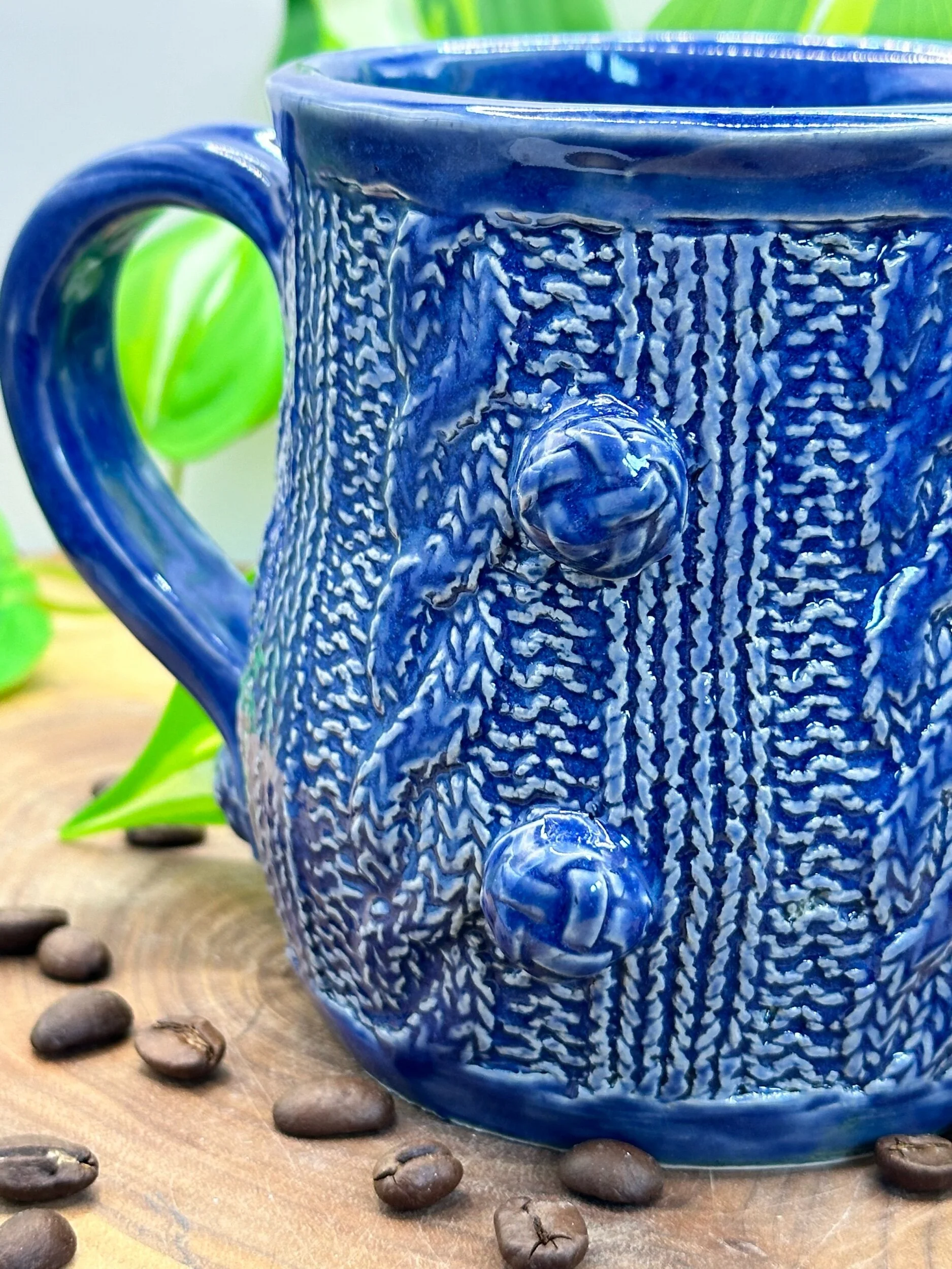 Close-up of a blue ceramic mug with textured patterns and decorative knobs, placed on a wooden surface with coffee beans and green leaves in the background.