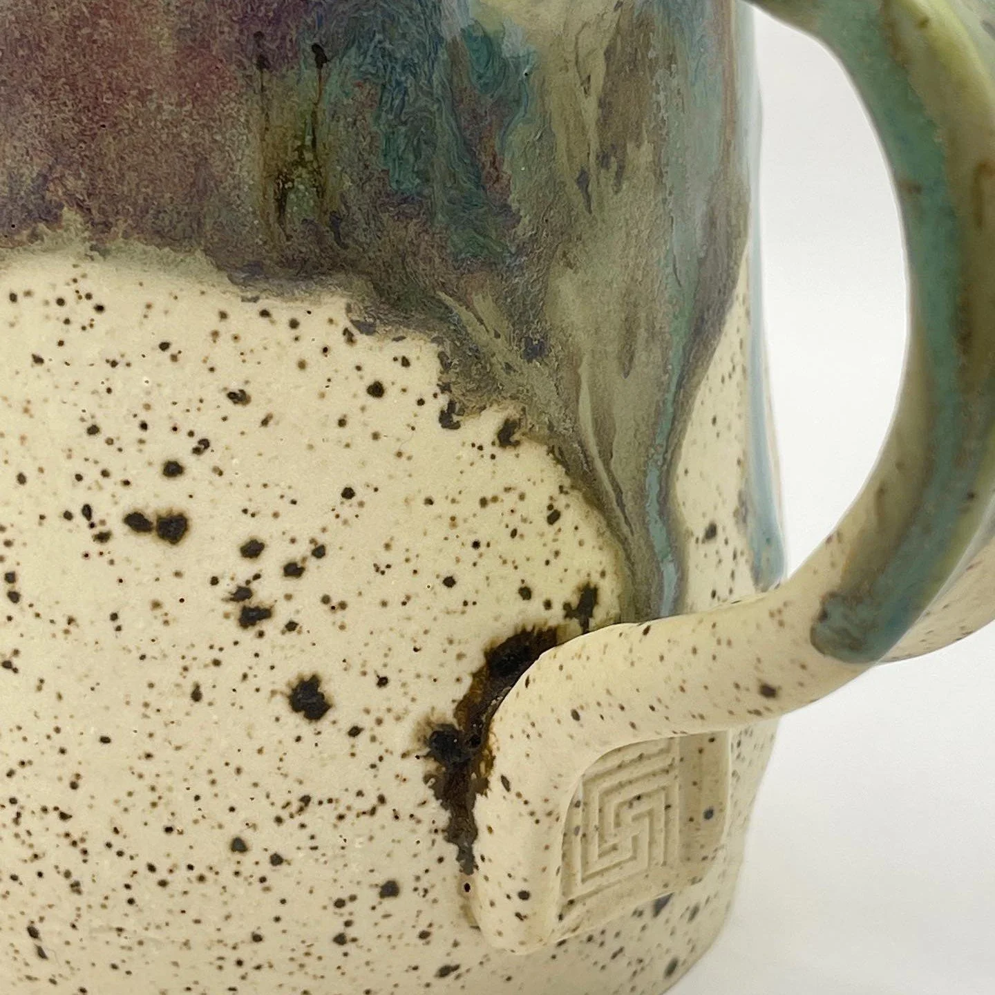Close-up of a textured ceramic mug with a speckled cream and black finish and a swirl of greenish-blue glaze near the rim and handle.