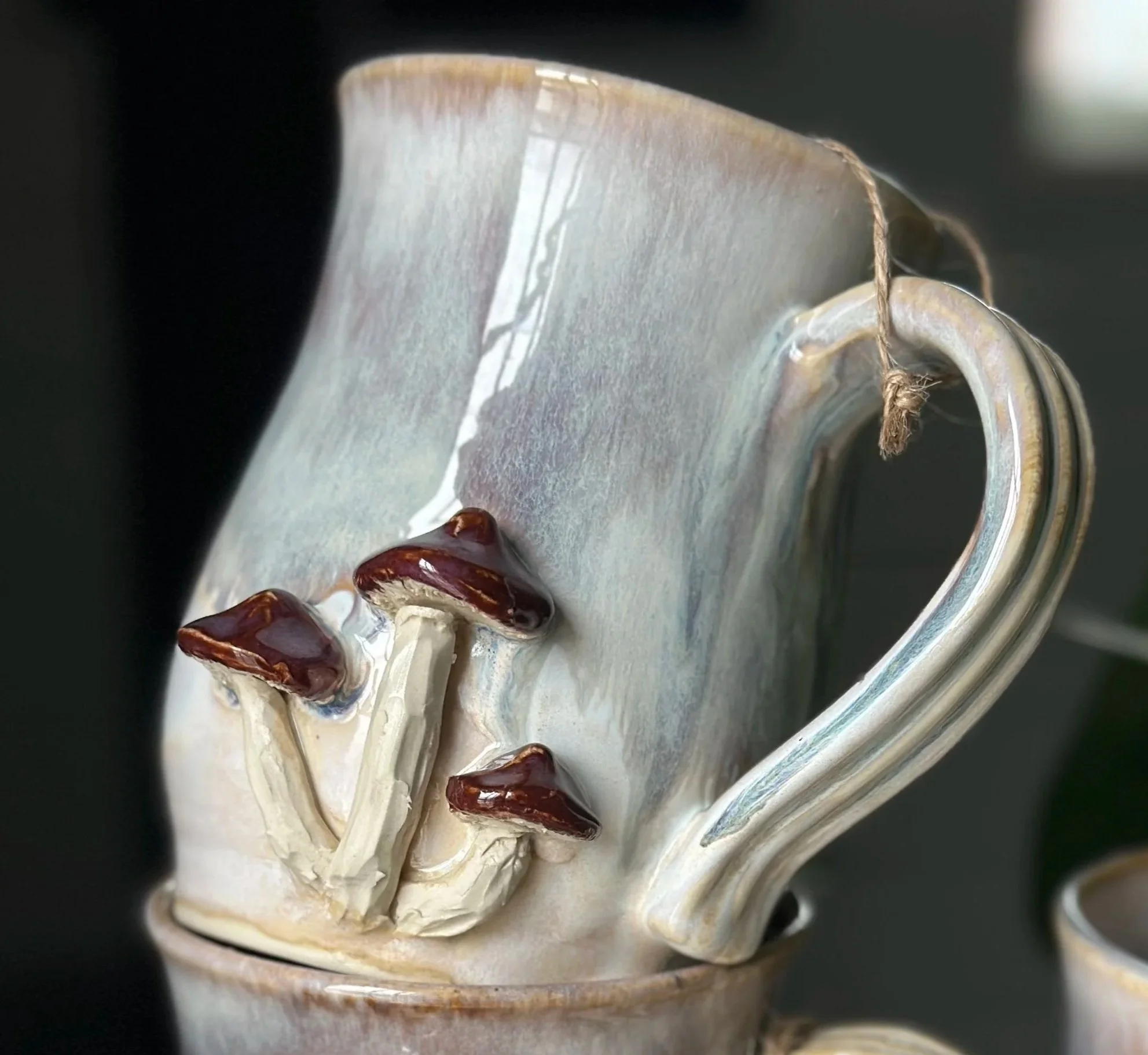 Close-up of a ceramic mug with mushroom-shaped accents on its surface, showing a smooth, glossy glaze and a hanging string on the rim.