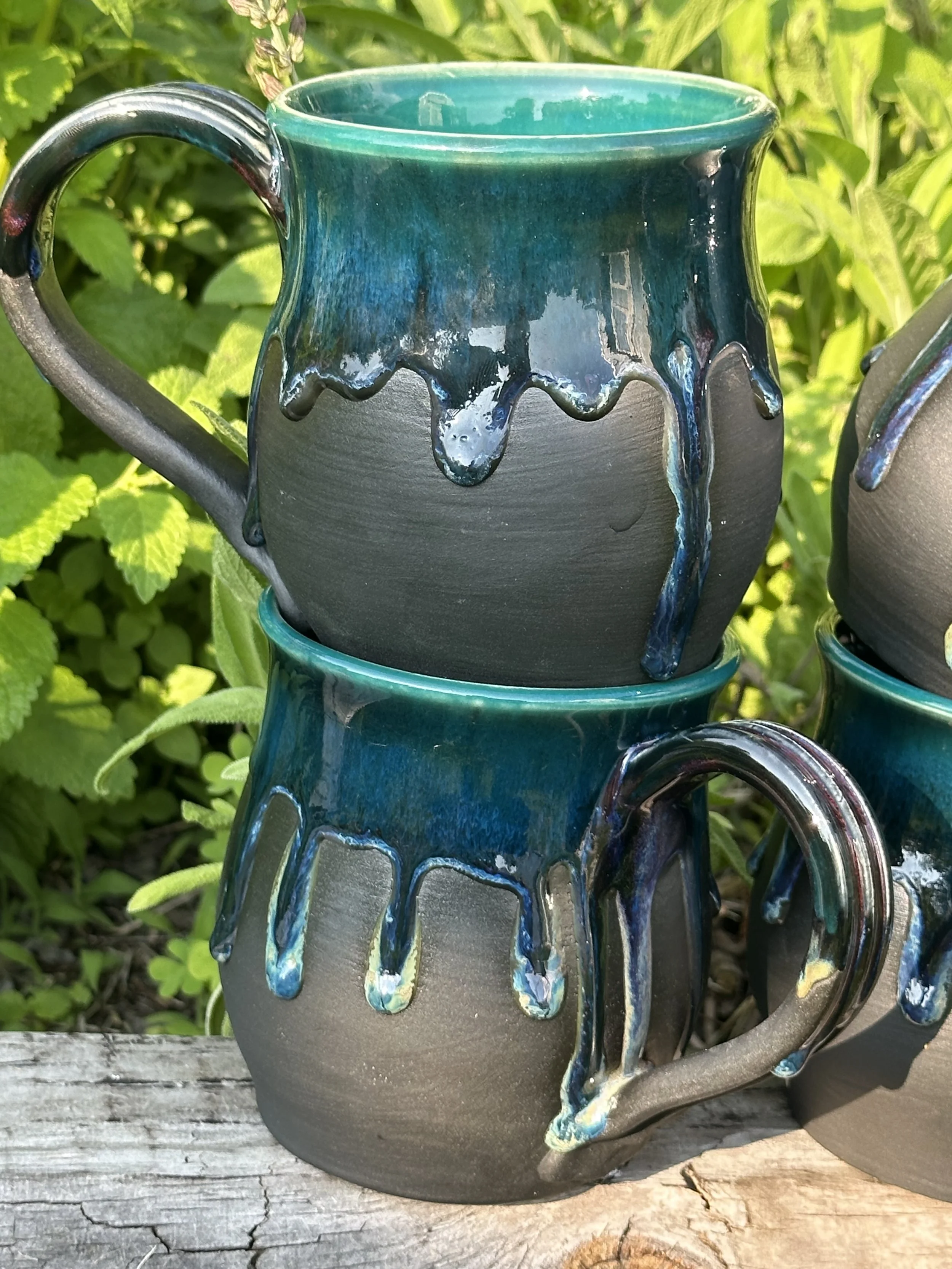 Two ceramic mugs with black and teal glaze, stacked outdoors on a weathered wooden surface, surrounded by green foliage.