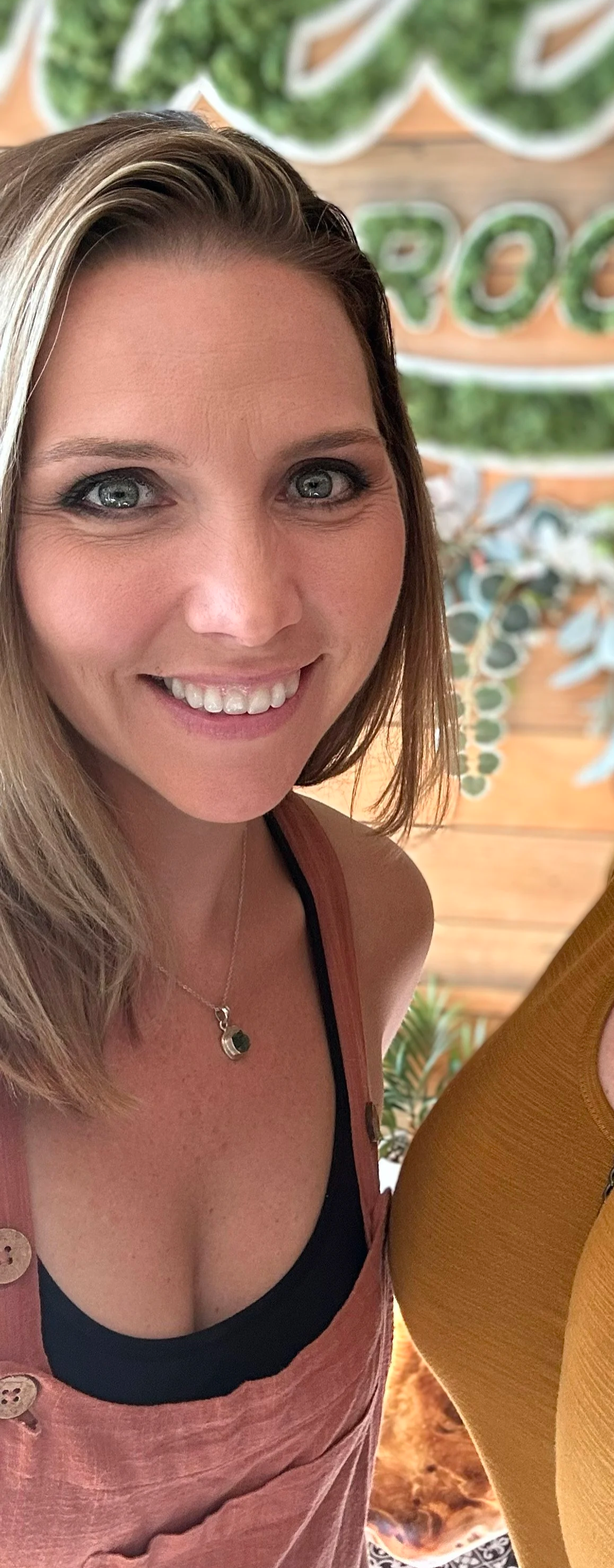 Close-up of a woman smiling, wearing a black top and a pink dress, with a necklace, in front of a blurred wooden background and greenery.
