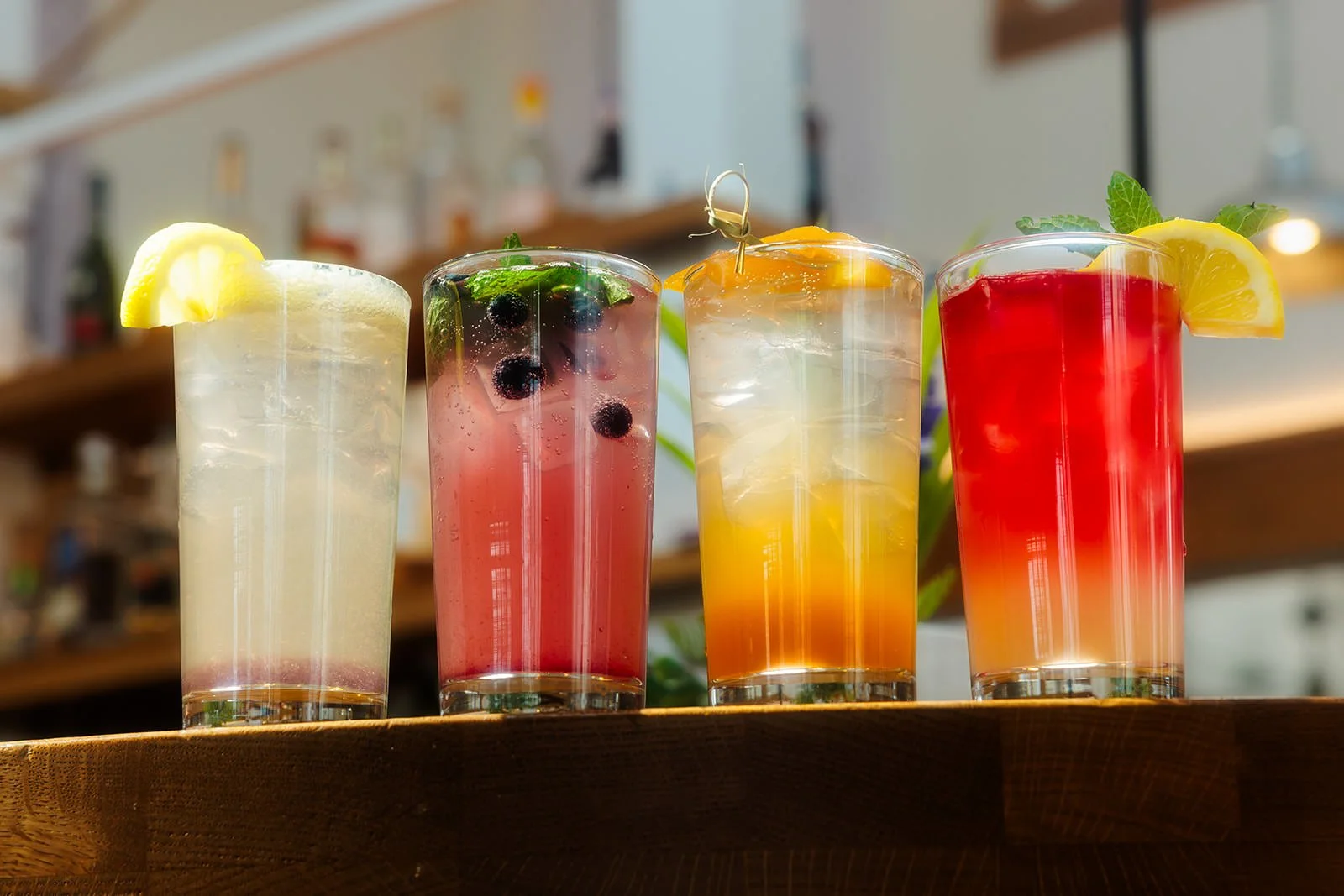 Four colorful glasses of different fruit drinks with lemon, blueberries, and mint garnishes on a wooden tray.