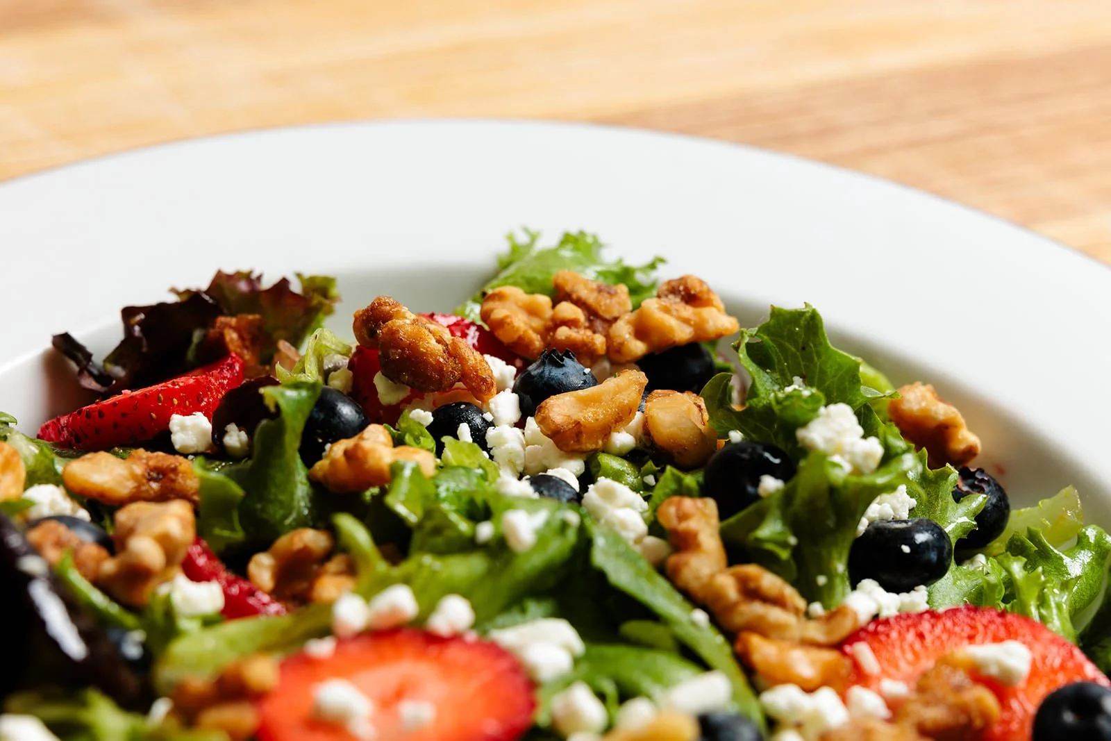 Fresh salad with strawberries, blueberries, walnuts, arugula, feta cheese, and greens in a white bowl on a wooden surface.