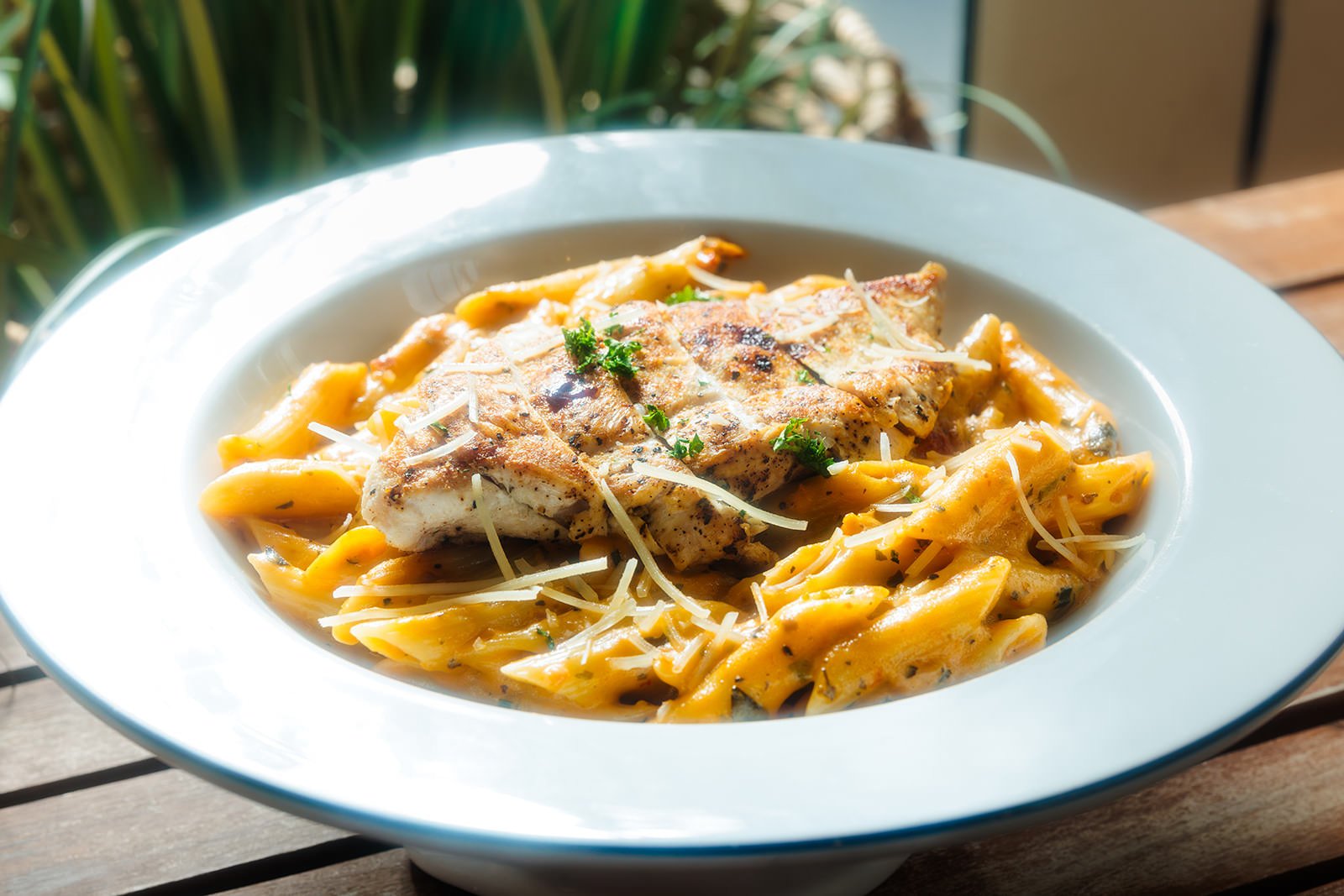 Plate of creamy fettuccine pasta with grilled chicken garnished with parsley and grated cheese, on a wooden table near a window.