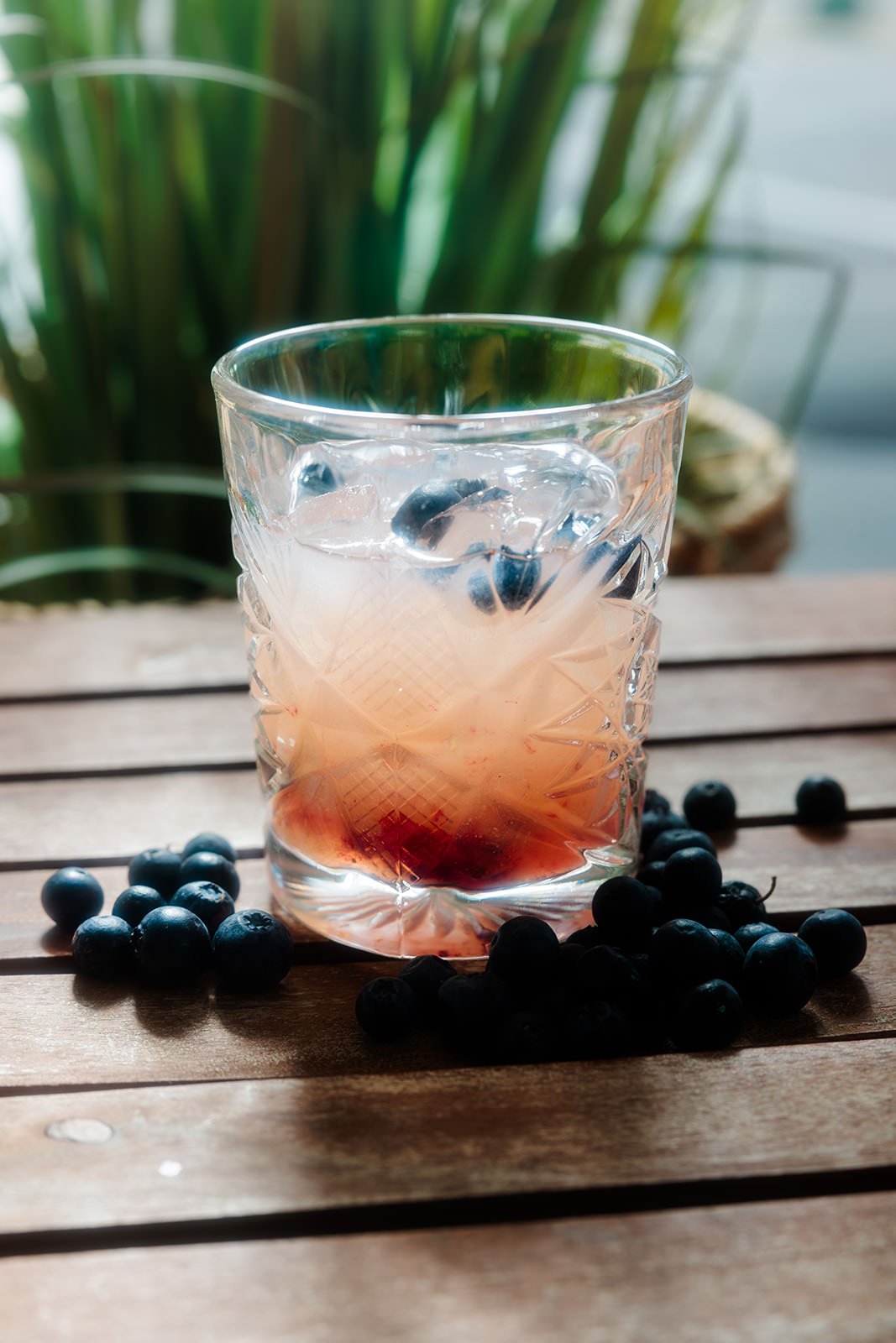 A glass tumbler with a textured pattern contains a pinkish beverage with ice and blueberries, placed on a wooden surface with scattered blueberries around it.