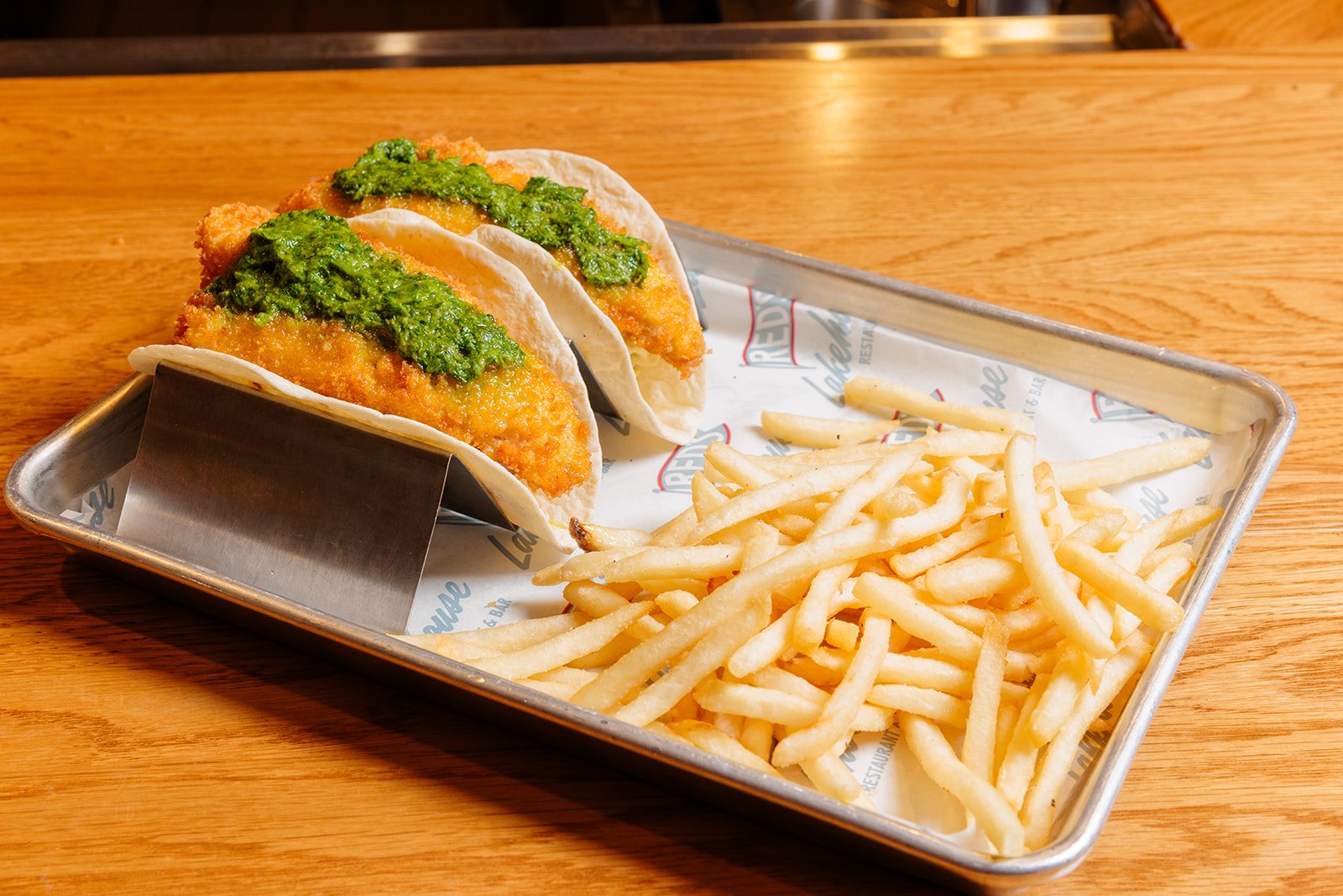 Two fish tacos with green sauce on a metal tray alongside French fries on a wooden table.
