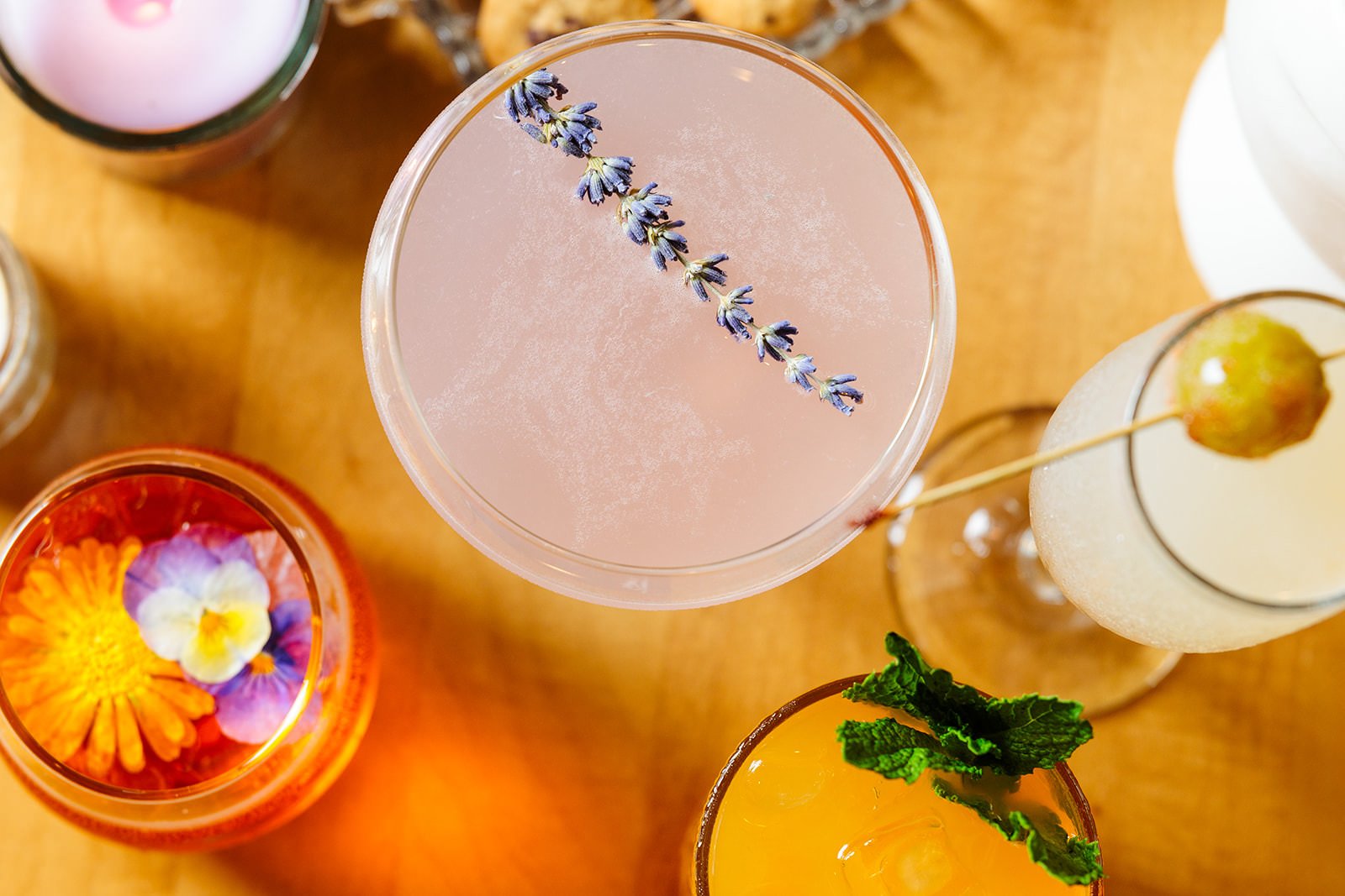 Top-down view of various colorful cocktails, one with a sprig of lavender, another with a rose and edible flowers, and others garnished with fruit and mint on a wooden surface.