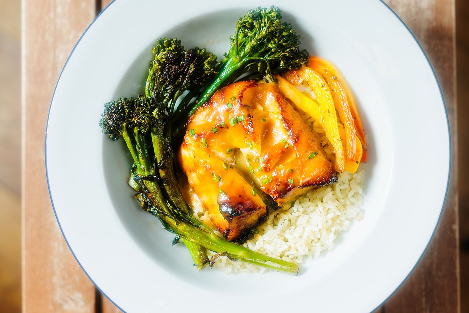 A plate of cooked white rice topped with a grilled fish fillet glazed with sauce, served with sautéed yellow and red bell peppers, and steamed broccolini.