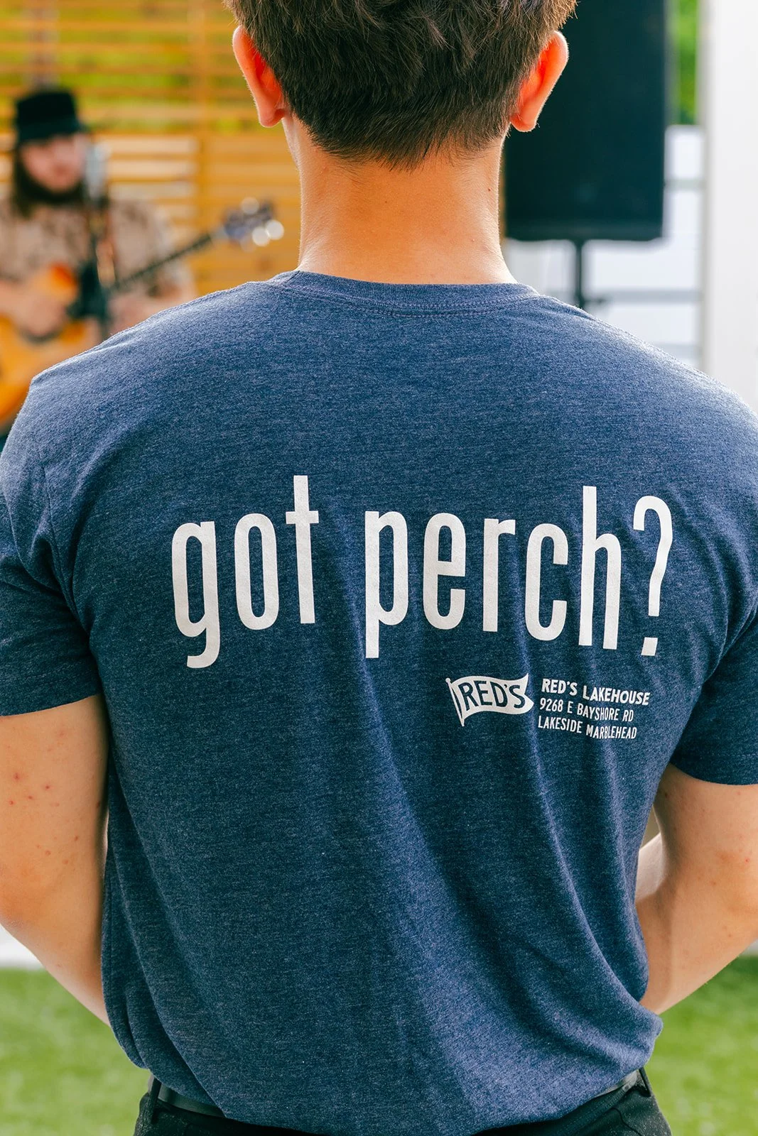 Back of a person wearing a blue T-shirt with white text that reads 'got perch?' and a logo for Red's Lakehouse with an address printed below.
