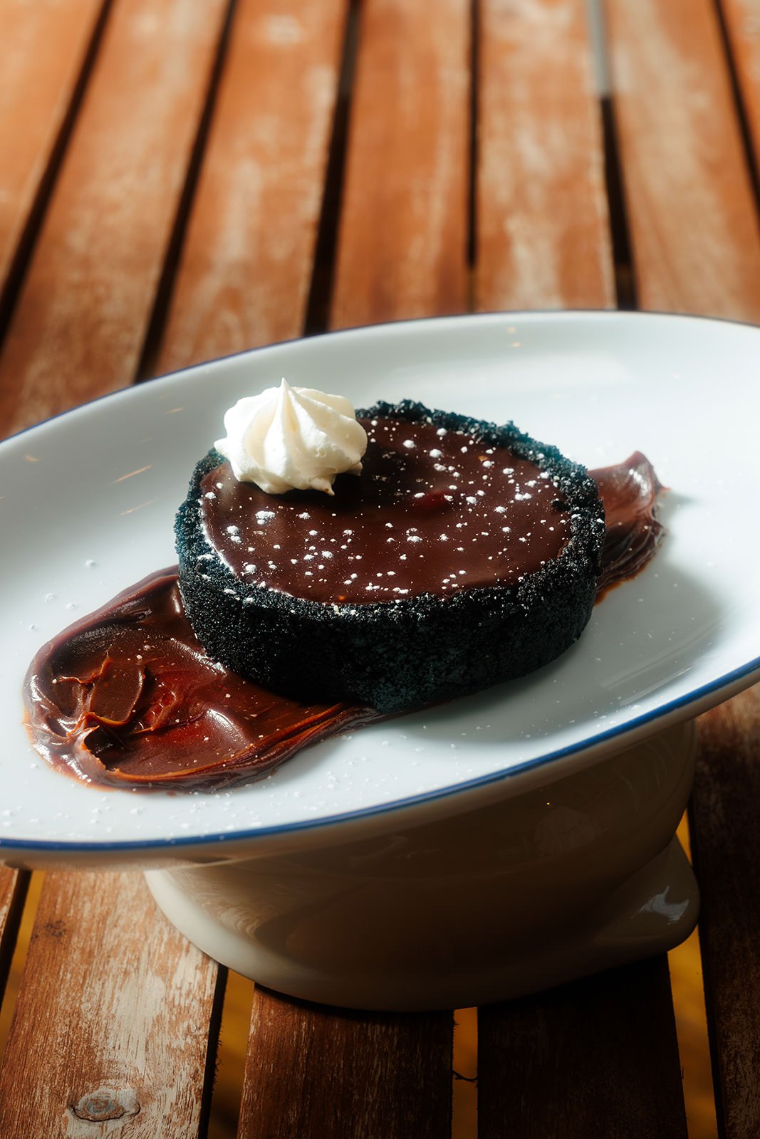 Gourmet dessert featuring a round chocolate cake with a dark, crusted exterior, topped with a dollop of whipped cream and sprinkled with powdered sugar, served on a white spoon-shaped dish with a chocolate smear underneath, on a wooden surface.