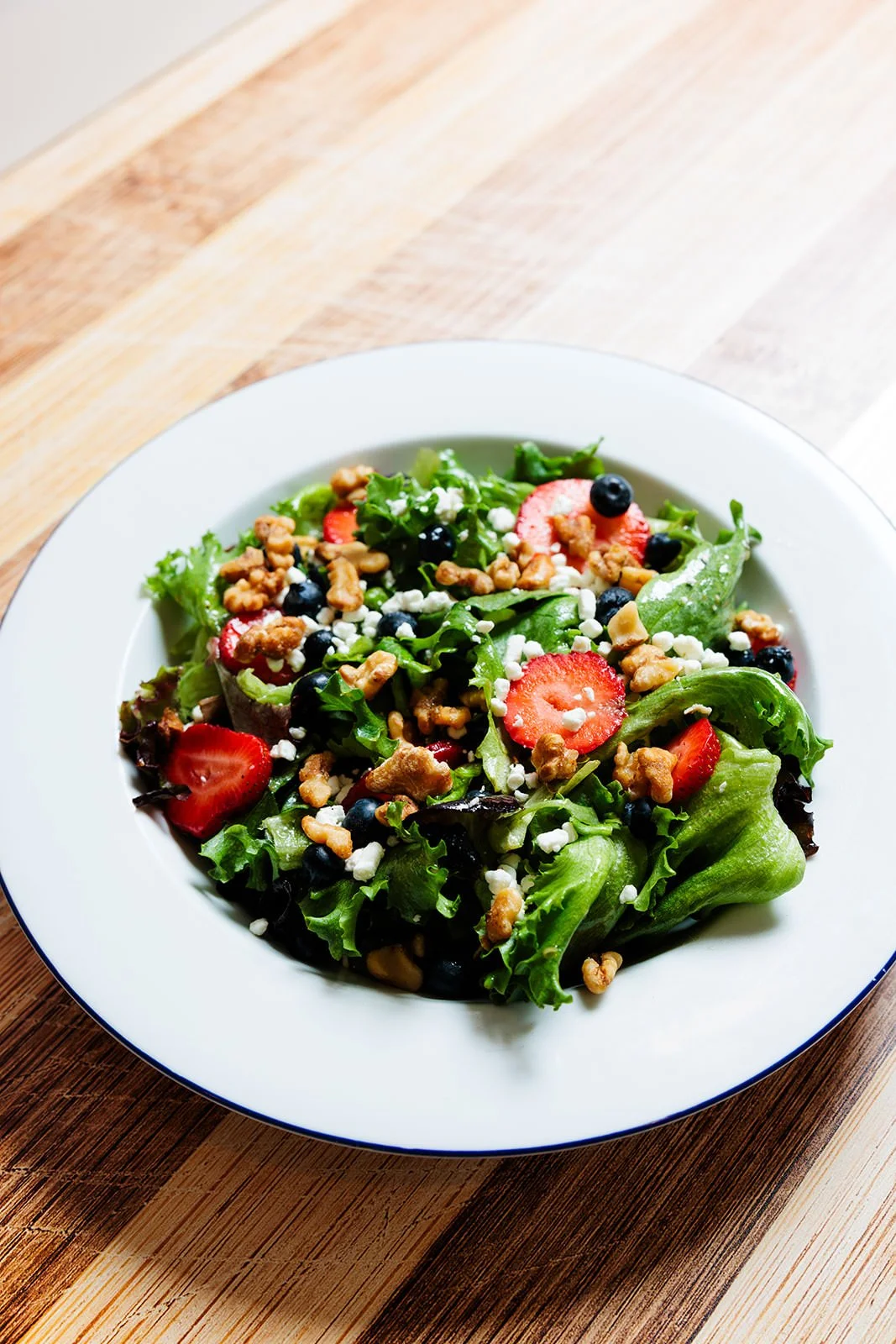 Fresh mixed green salad with strawberries, blueberries, crumbled cheese, and walnuts on a white plate.
