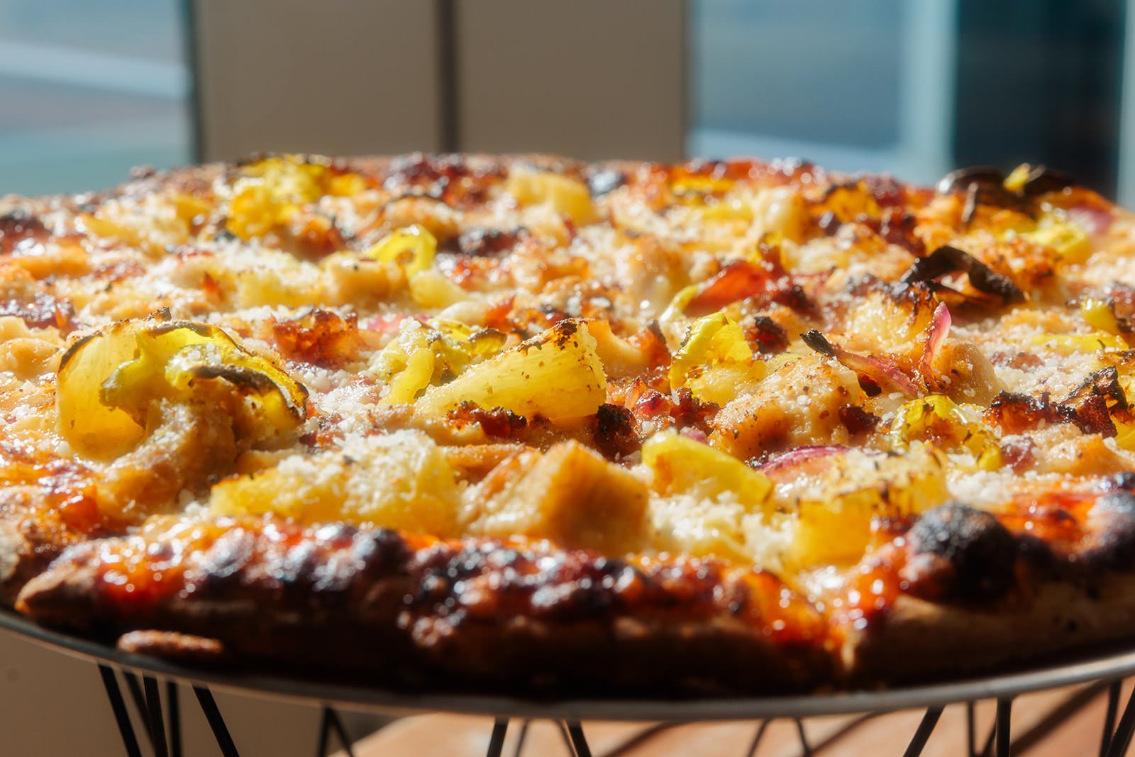 Close-up of a pizza with pineapple chunks, bacon, cheese, and other toppings on a black wire pizza rack.