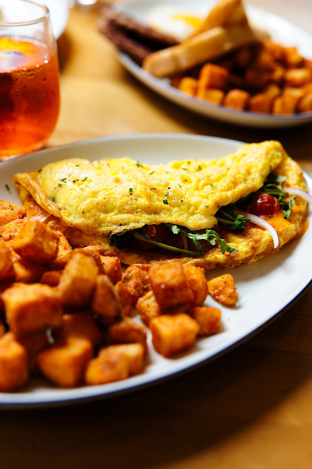 Close-up of a breakfast plate with scrambled eggs, roasted potatoes, and an omelet with vegetables, alongside a glass of iced tea, with a second plate of toast and bacon in the background.