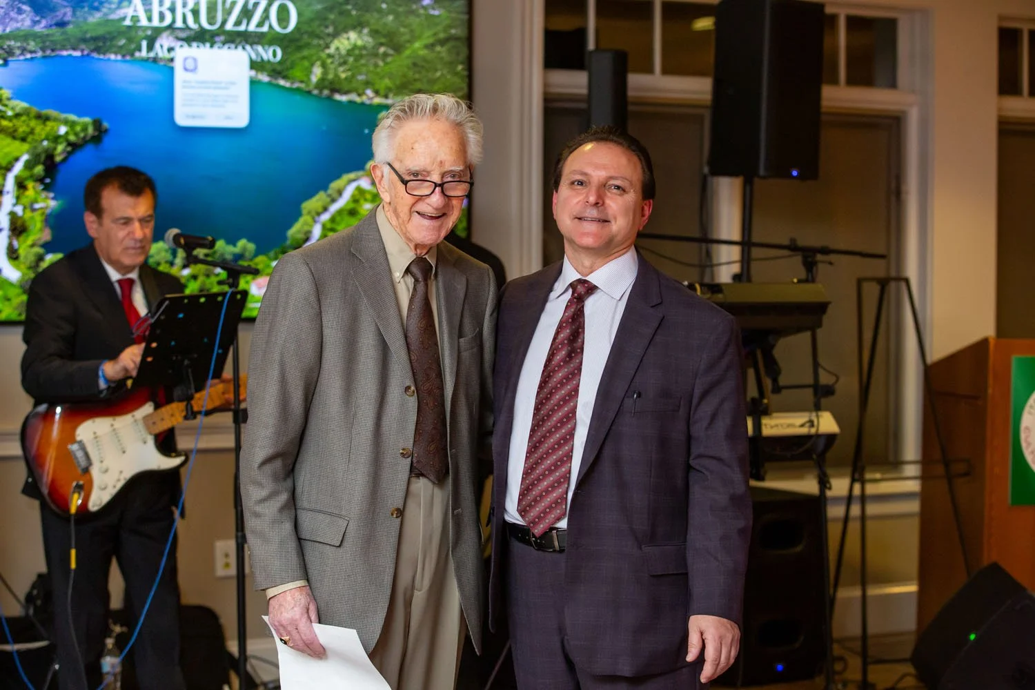 Two men in suits posing for a photo at an indoor event, with a musician playing guitar in the background and a large screen displaying a lake and greenery.