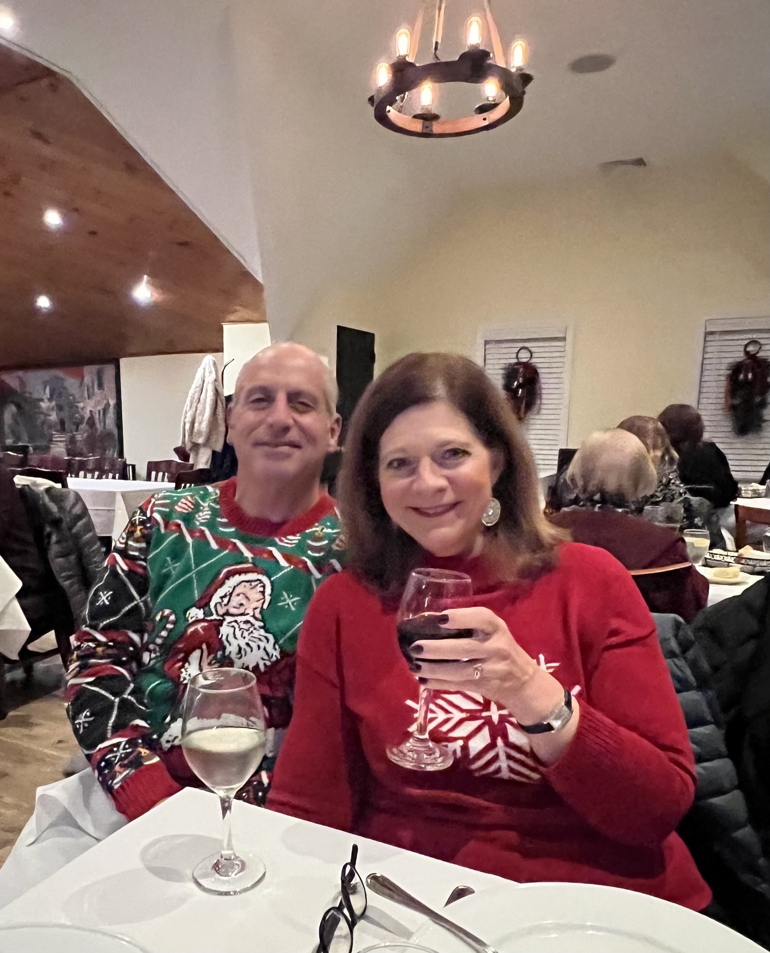 A couple sitting at a restaurant table, wearing festive Christmas sweaters. The man is in a Santa-themed sweater, and the woman is in a red sweater with a snowflake design. Both are holding wine glasses. A chandelier hangs above, and diners fill the 