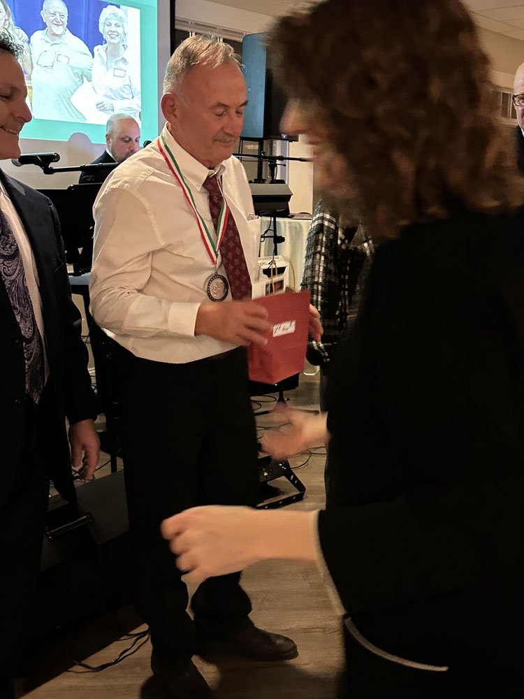 A man wearing a medal around his neck, holding a red gift bag, at an indoor event with people and a screen in the background.