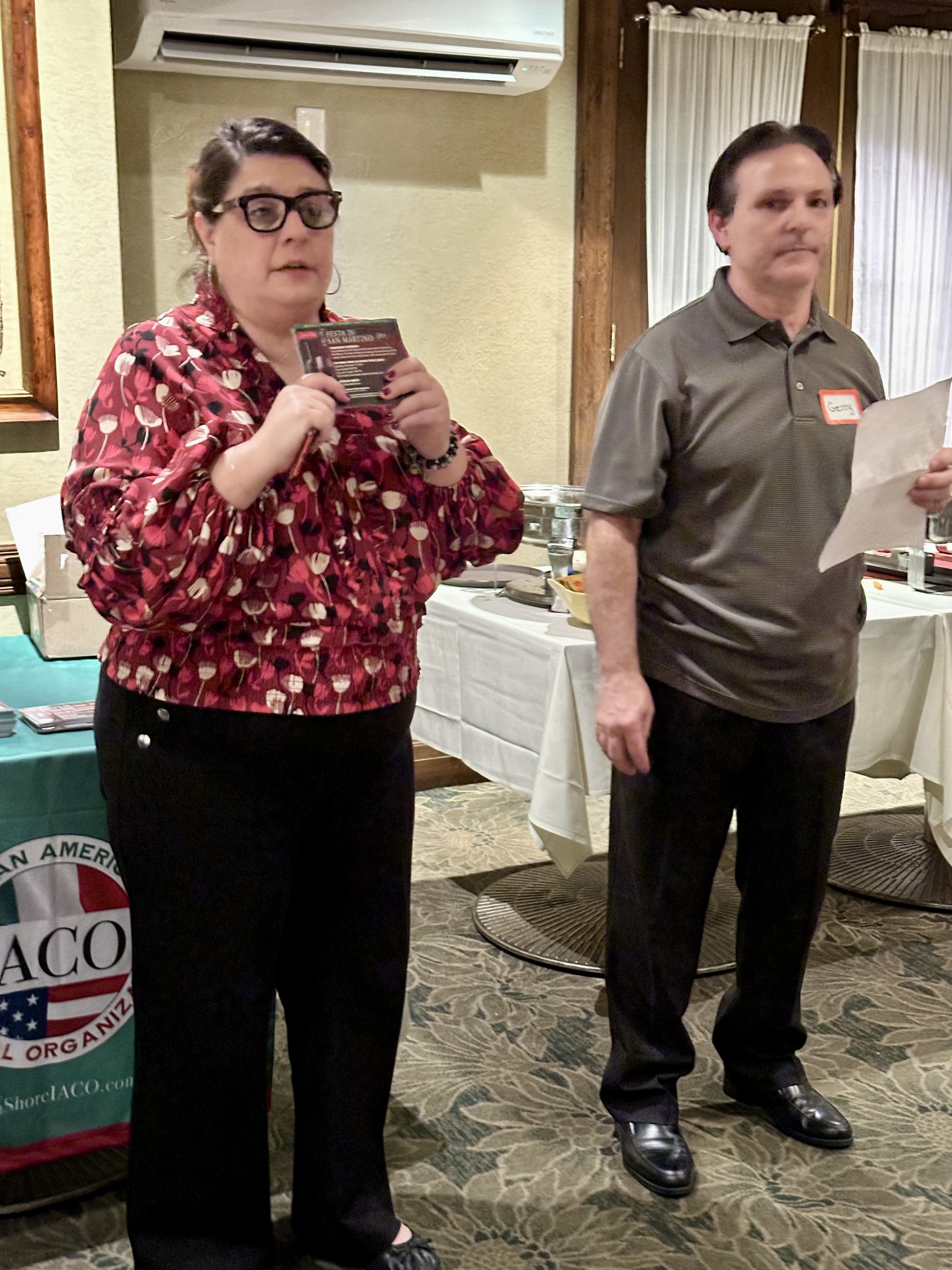 Two people standing indoors, one holding a flyer and the other holding a paper.