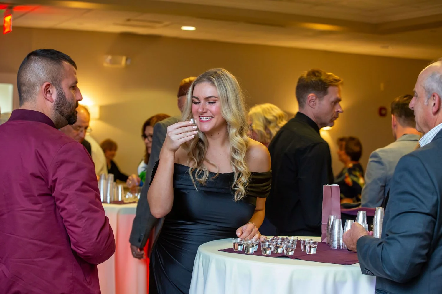 People socializing at a cocktail party, with a woman in a black dress laughing and holding a drink, and men in formal attire standing around a high table with shot glasses and cups.