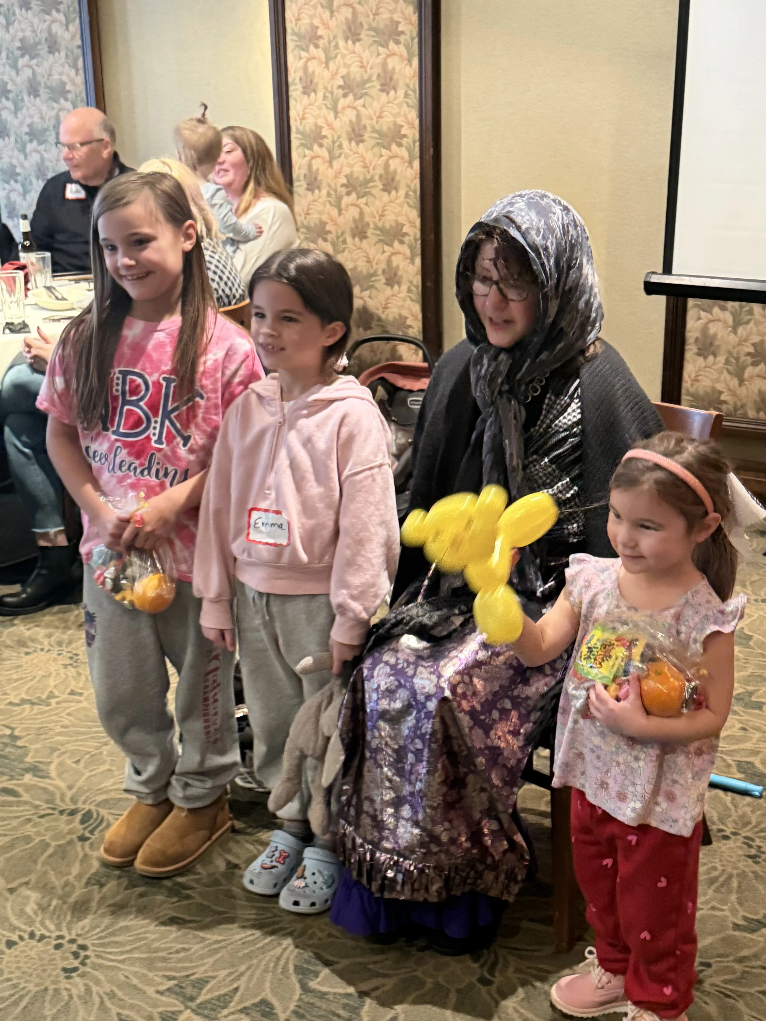 A group of children and an older woman at an indoor gathering. The children are holding bags of snacks, and one girl is holding yellow balloons. The older woman is sitting and holding the balloons. Others are visible in the background, seated at a ta