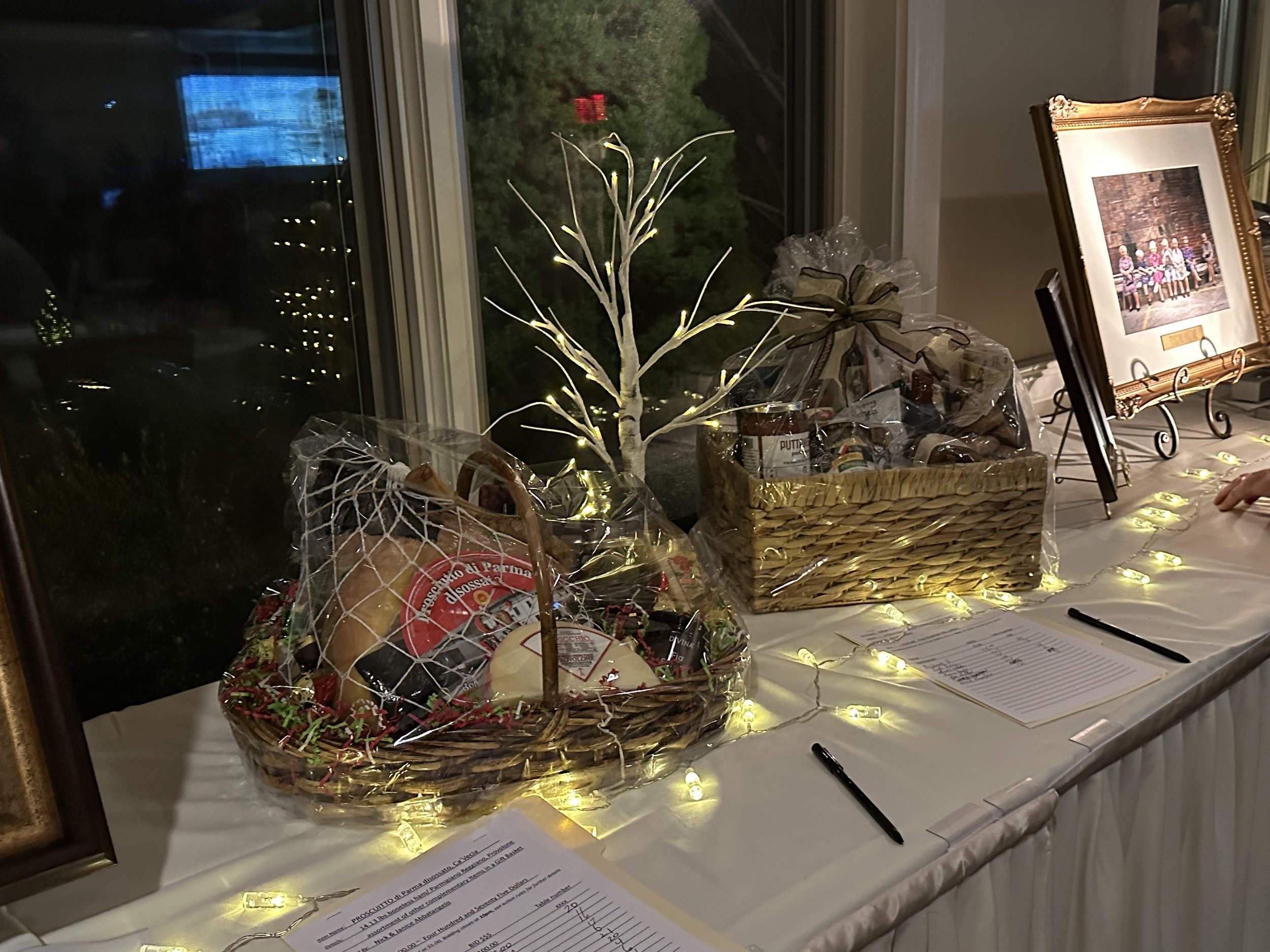 A table display featuring gift baskets wrapped in cellophane, a framed picture on an easel, decorative string lights, and pens on bid sheets for a silent auction.